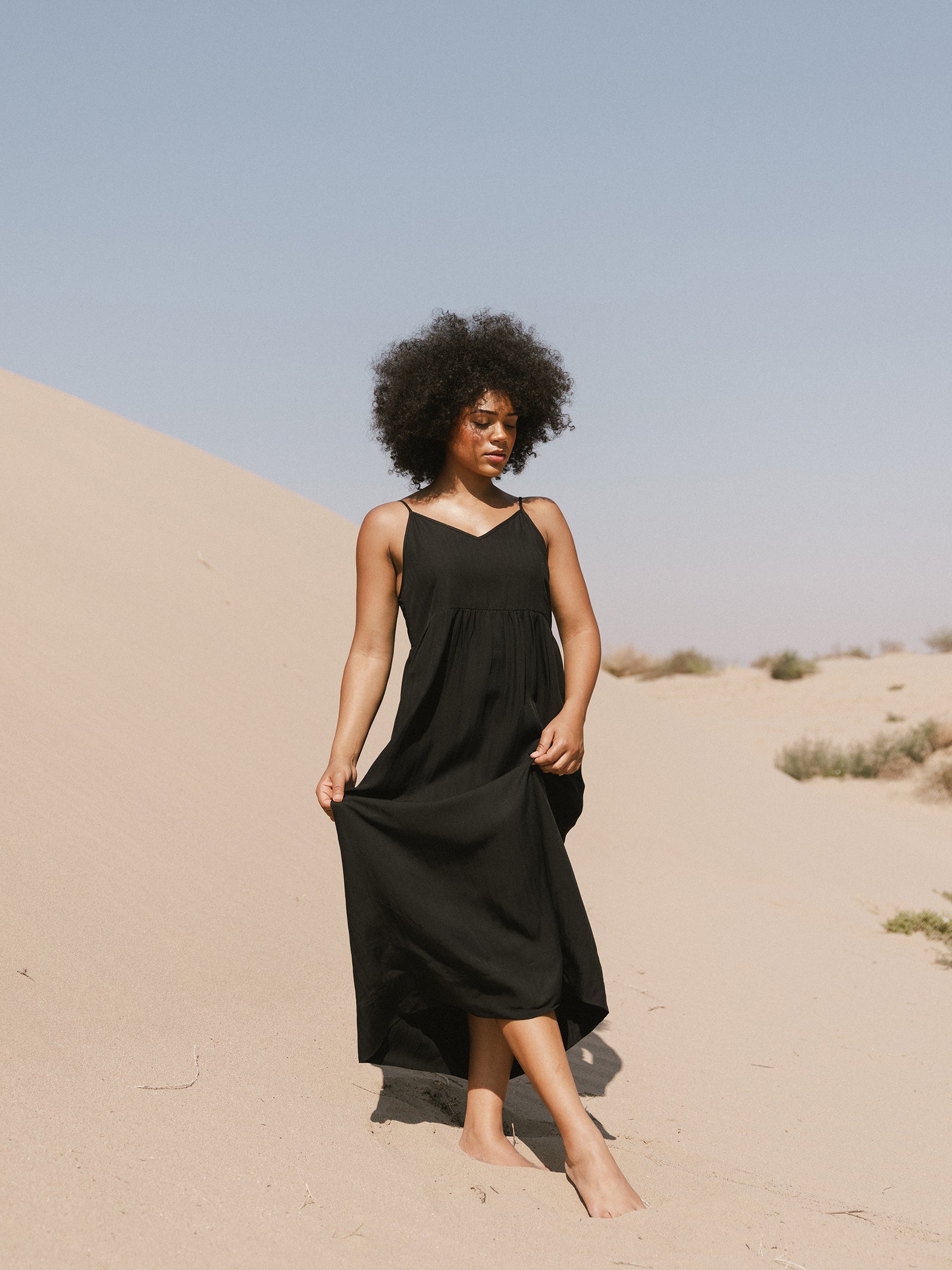 A person with curly hair stands barefoot on a sandy landscape, wearing Cozy Earth's Women's Sunset Gathered Dress. They hold the hem of the sleeveless black dress with one hand and look away from the camera. The sky is clear and blue. 