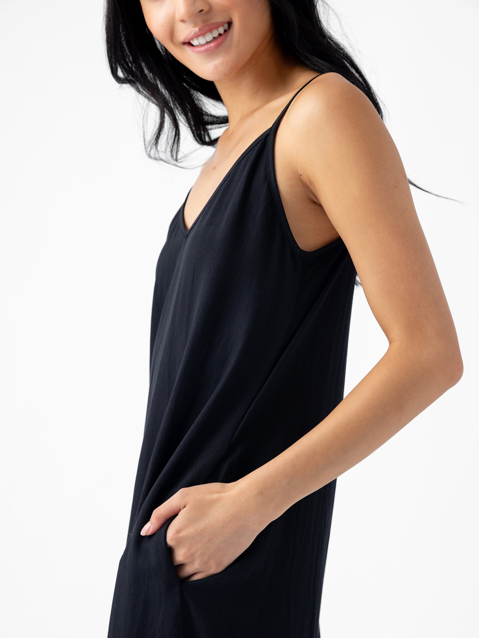 A person with long black hair smiles and wears the Women's Sunset Slip Dress by Cozy Earth. The person has one hand in a pocket, and the background is white. 