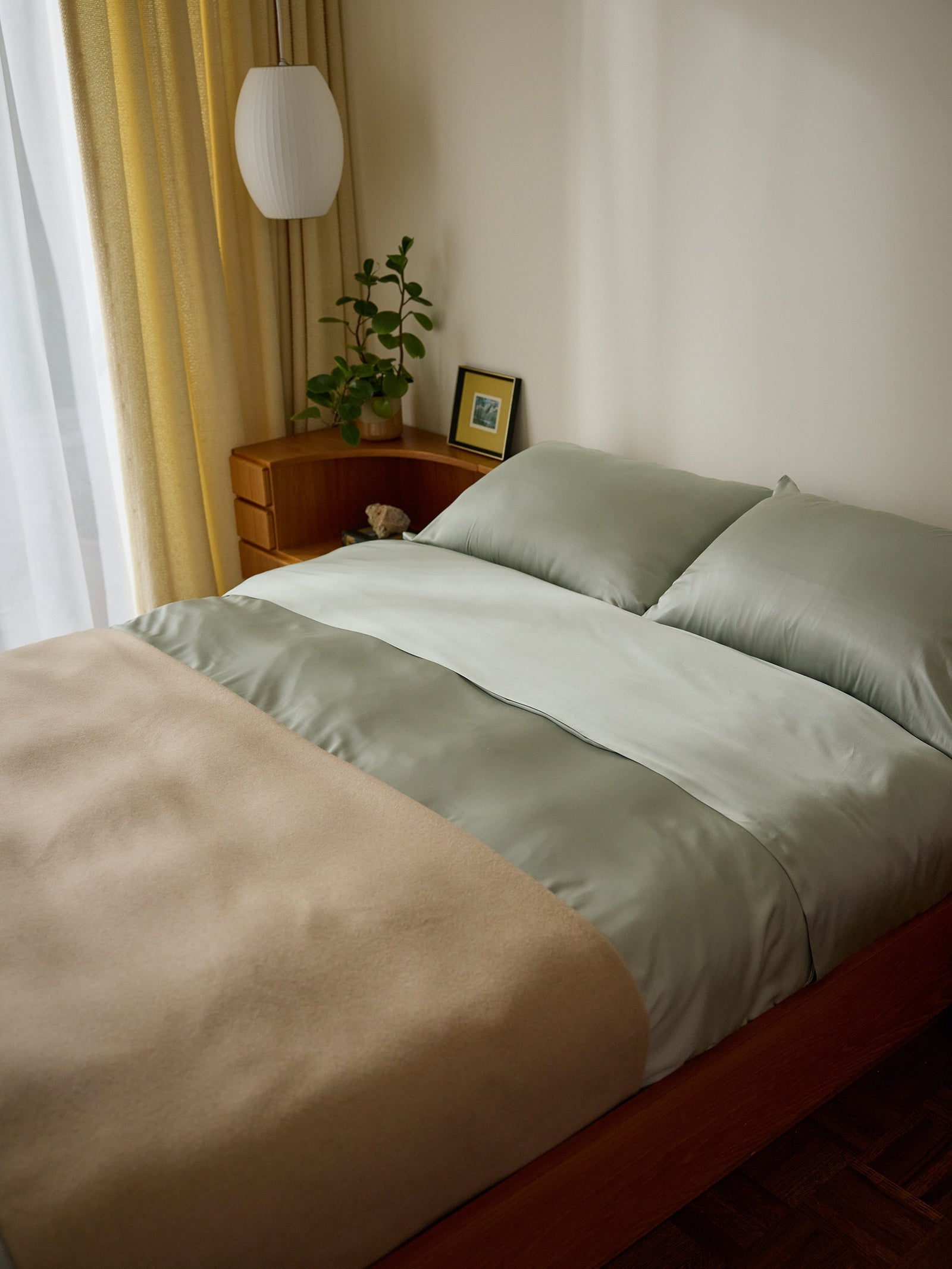 A neatly made bed featuring the Cozy Earth Bamboo Duvet Cover in light green and a beige blanket sits in a cozy bedroom. A wooden nightstand has a small plant, framed picture, and white hanging lamp, while yellow curtains allow natural light from the window. 