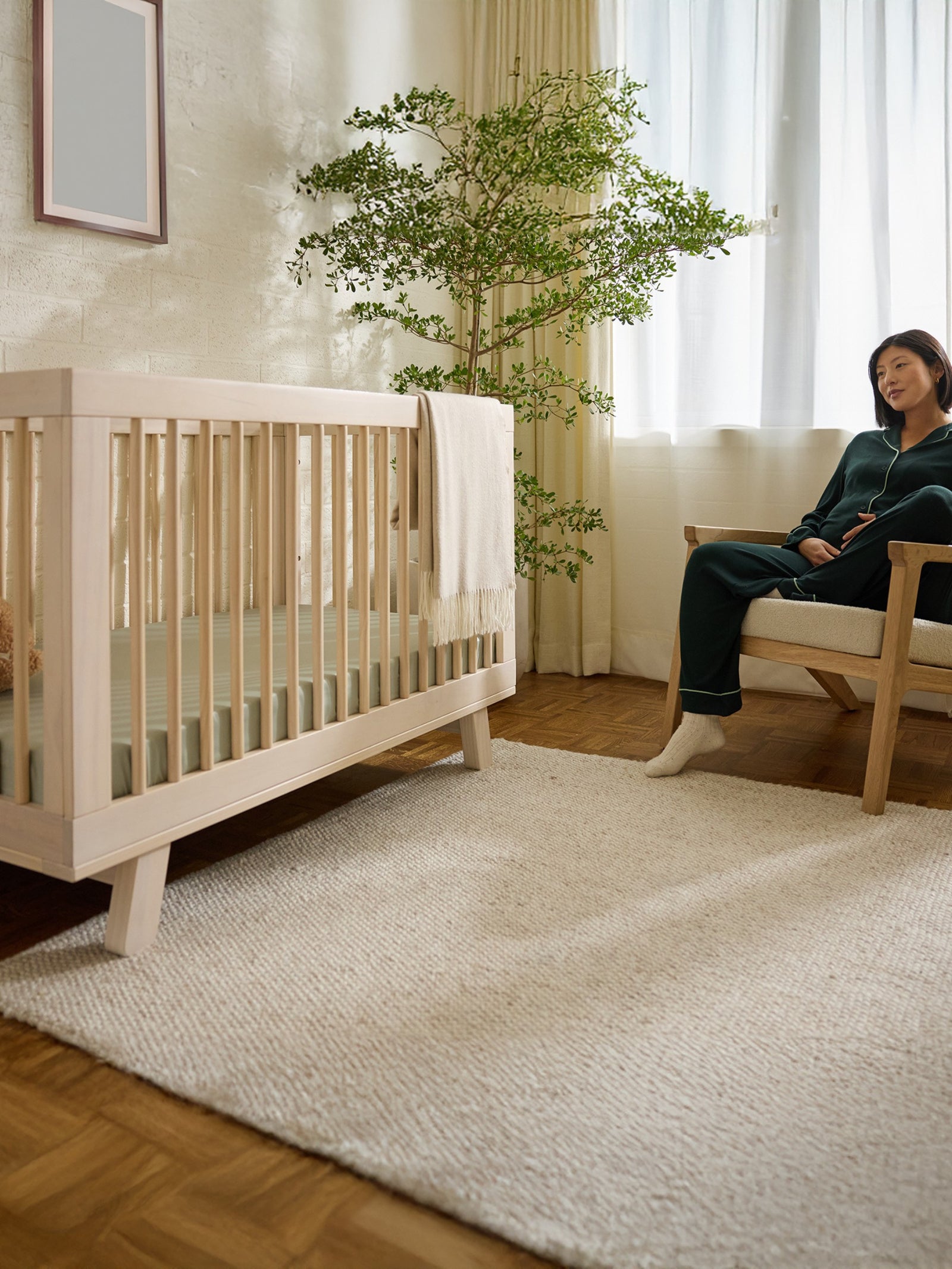 A woman sits beside a wooden crib adorned with a Cozy Earth Crib Sheet in a softly lit nursery. A blanket drapes over the crib, and a large plant stands nearby as light streams through the window, creating a calm atmosphere. 