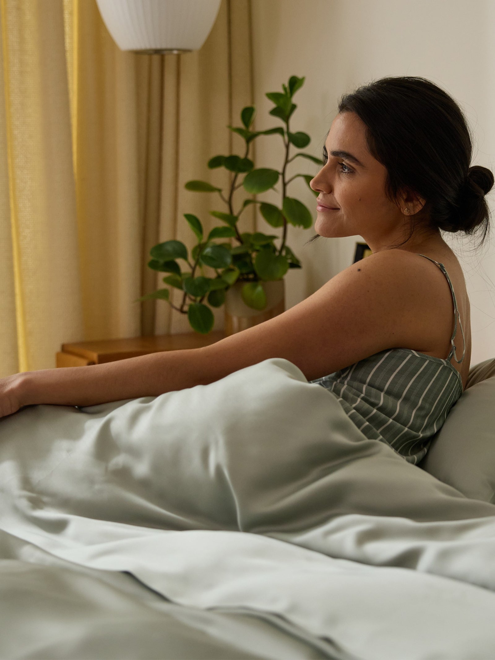 A woman in a striped nightgown sits up in bed, wrapped in a Cozy Earth Bamboo Flat Sheet. She gazes to the side with a serene expression, while a plant and lamp softly lit in the background enhance the cozy atmosphere. 