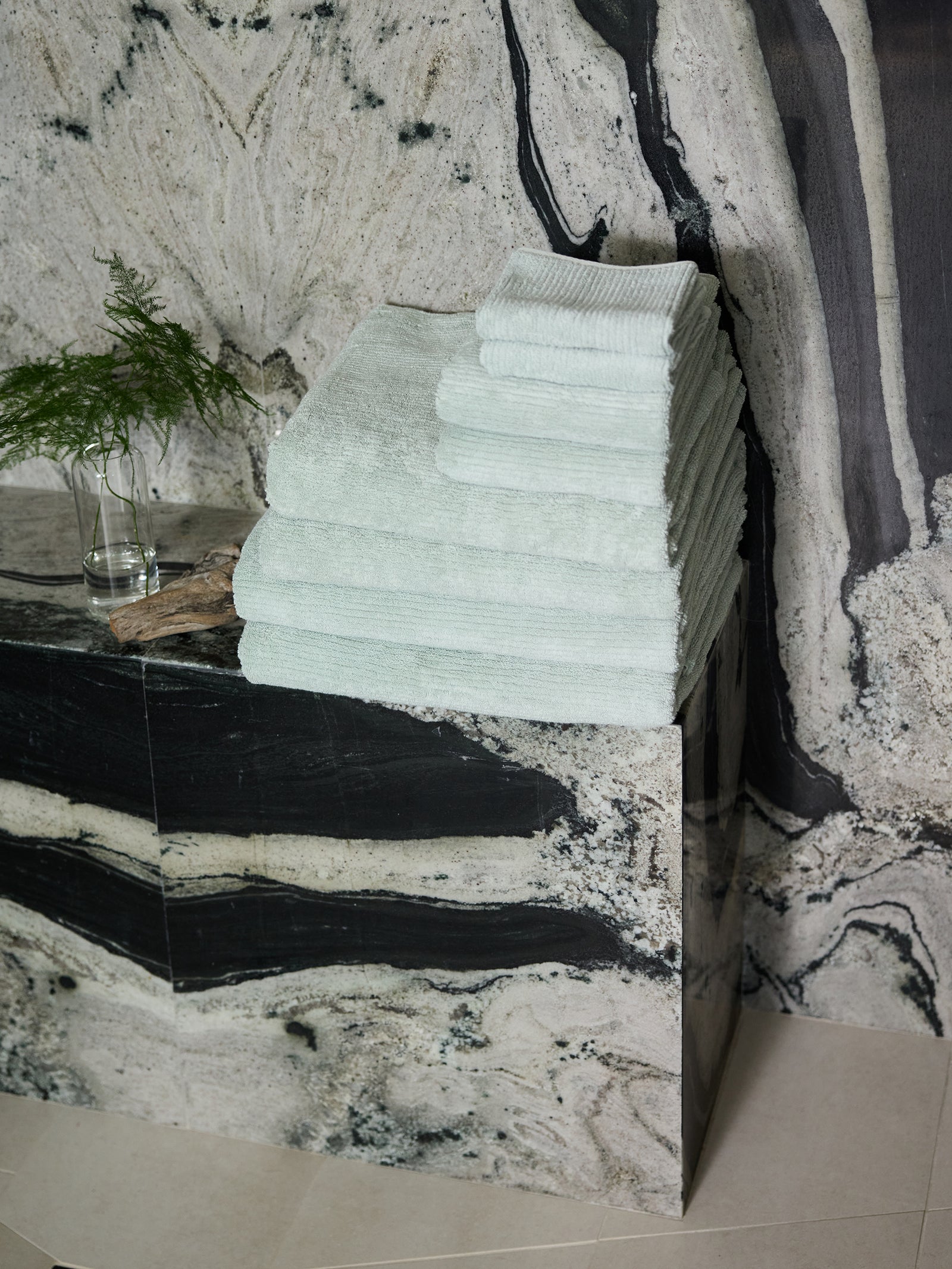 A stack of Cozy Earth's Ribbed Terry Bath Towel Set, with its pale green hue, is neatly arranged on a marble shelf. Nearby, a small vase holds green foliage against the backdrop of an elegant marbled wall featuring shades of black, gray, and white. 