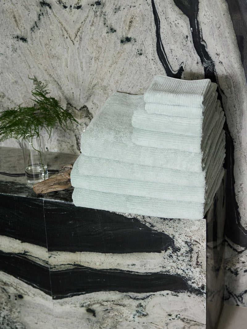 The Complete Ribbed Terry Bath Bundle by Cozy Earth is stacked neatly on a black and white marble counter beside a vase with a green fern, set against a matching marble wall.