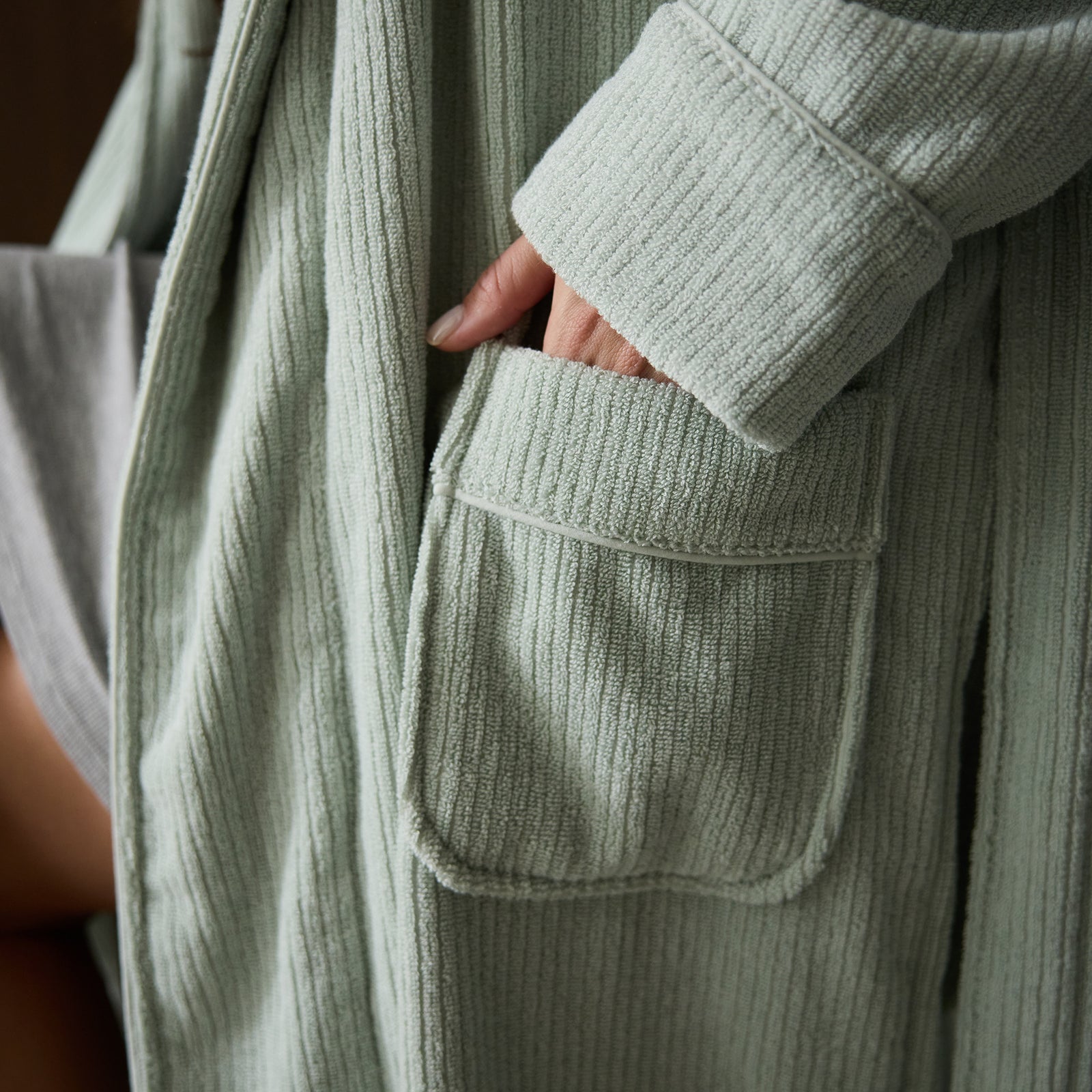 Close-up of a person wearing a light green Ribbed Terry Bath Robe by Cozy Earth, with their right hand tucked into a front pocket. The texture of the fabric appears soft and cozy. A glimpse of their grey inner garment is visible. 