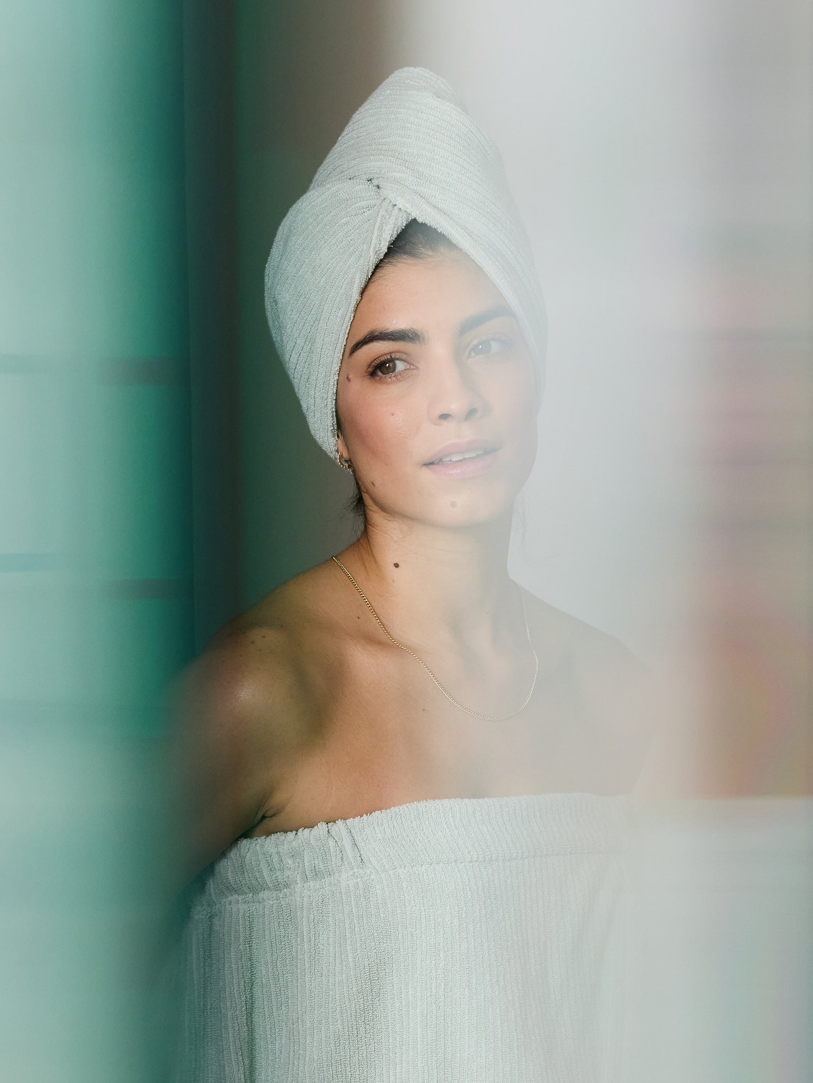 A woman with a Ribbed Terry Hair Towel from Cozy Earth wrapped around her head and a matching towel dress stands in a softly lit room. She gazes slightly upward with a serene expression, while the image showcases a soft-focus effect accompanied by subtle blueish-green tones. 