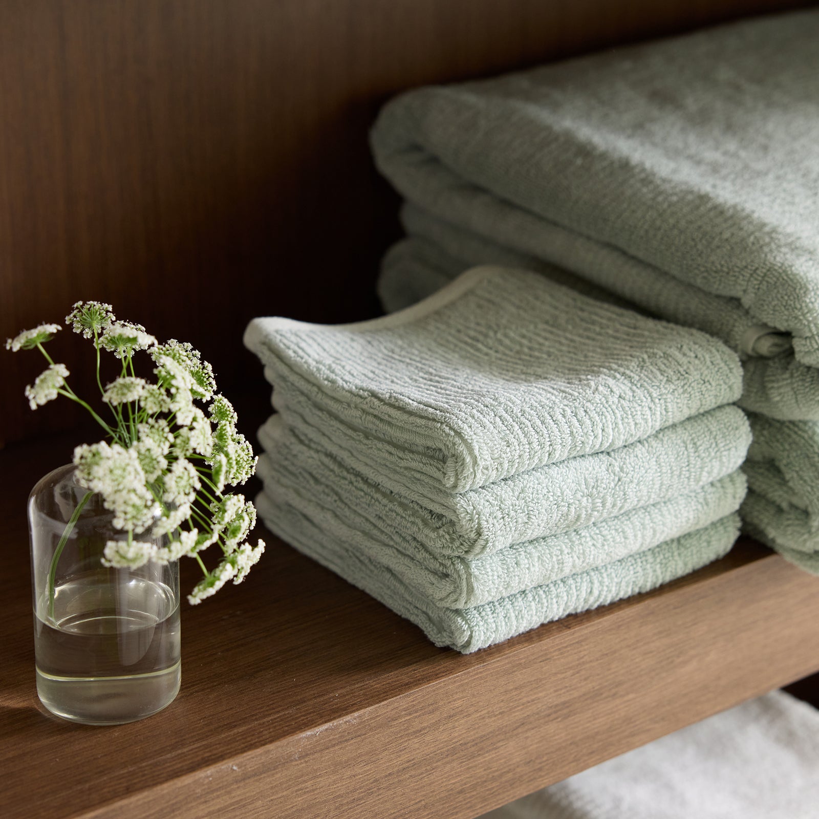 A neatly folded stack of Cozy Earth's Ribbed Terry Washcloths in pale green is placed on a wooden shelf. Beside the washcloths, a small clear glass with a few delicate white flowers in water adds a touch of elegance to the display. 