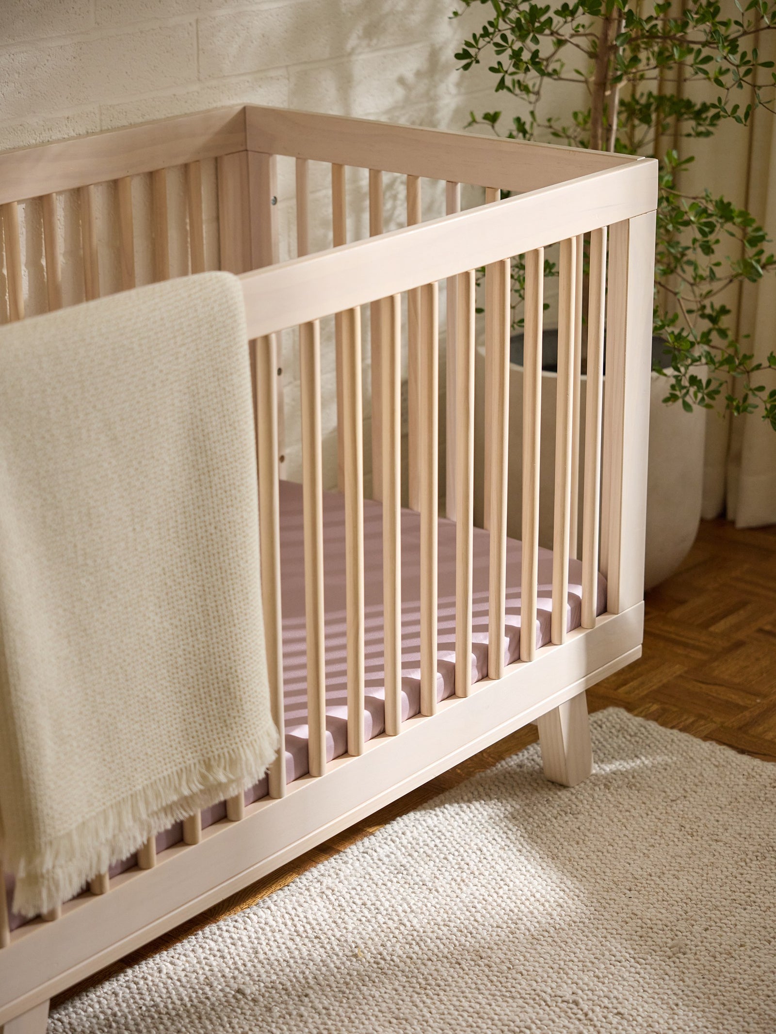 A Cozy Earth Crib Sheet from Cozy Earth adorns a wooden baby crib with a cream knitted blanket draped over one side, standing on a woven rug. A potted plant sits in the corner with sunlight filtering through nearby curtains. 