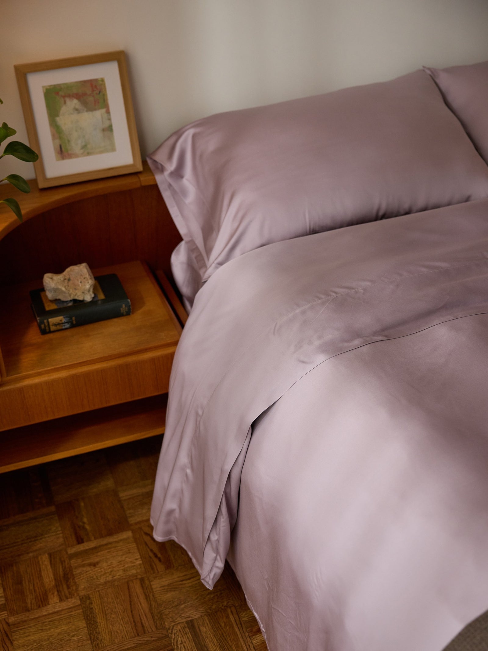 A neatly made bed with Cozy Earth's Bamboo Pillowcases in lavender is beside a wooden nightstand. The nightstand features a plant, framed picture, decorative stone, and book on its surface. A parquet floor lies in the foreground. 