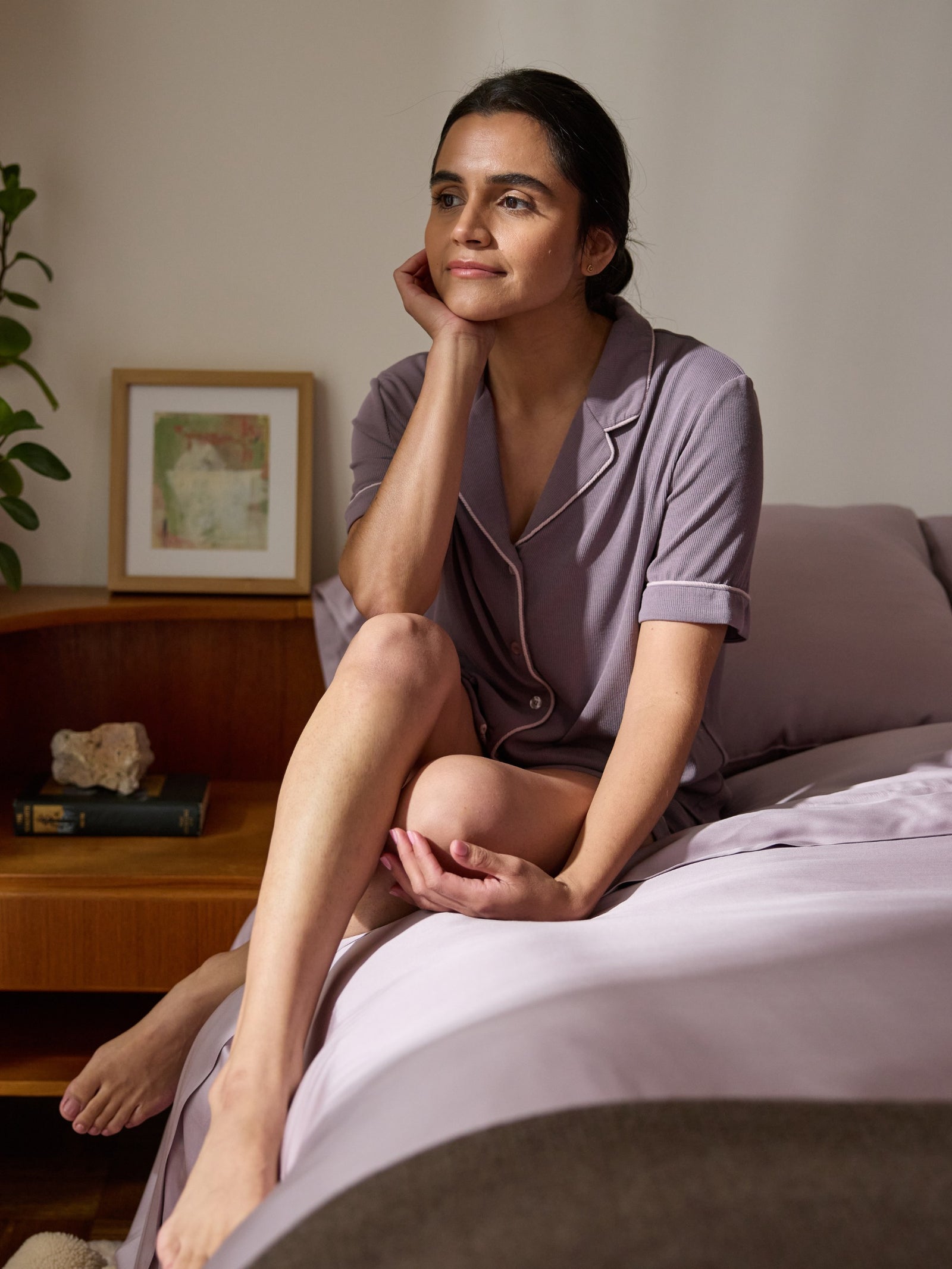 A person in lavender pajamas sits thoughtfully on a bed, one leg crossed. In the background are a plant and framed artwork beside a neatly made bed adorned with Cozy Earth's Bamboo Sheet Set and matching lavender duvet. 