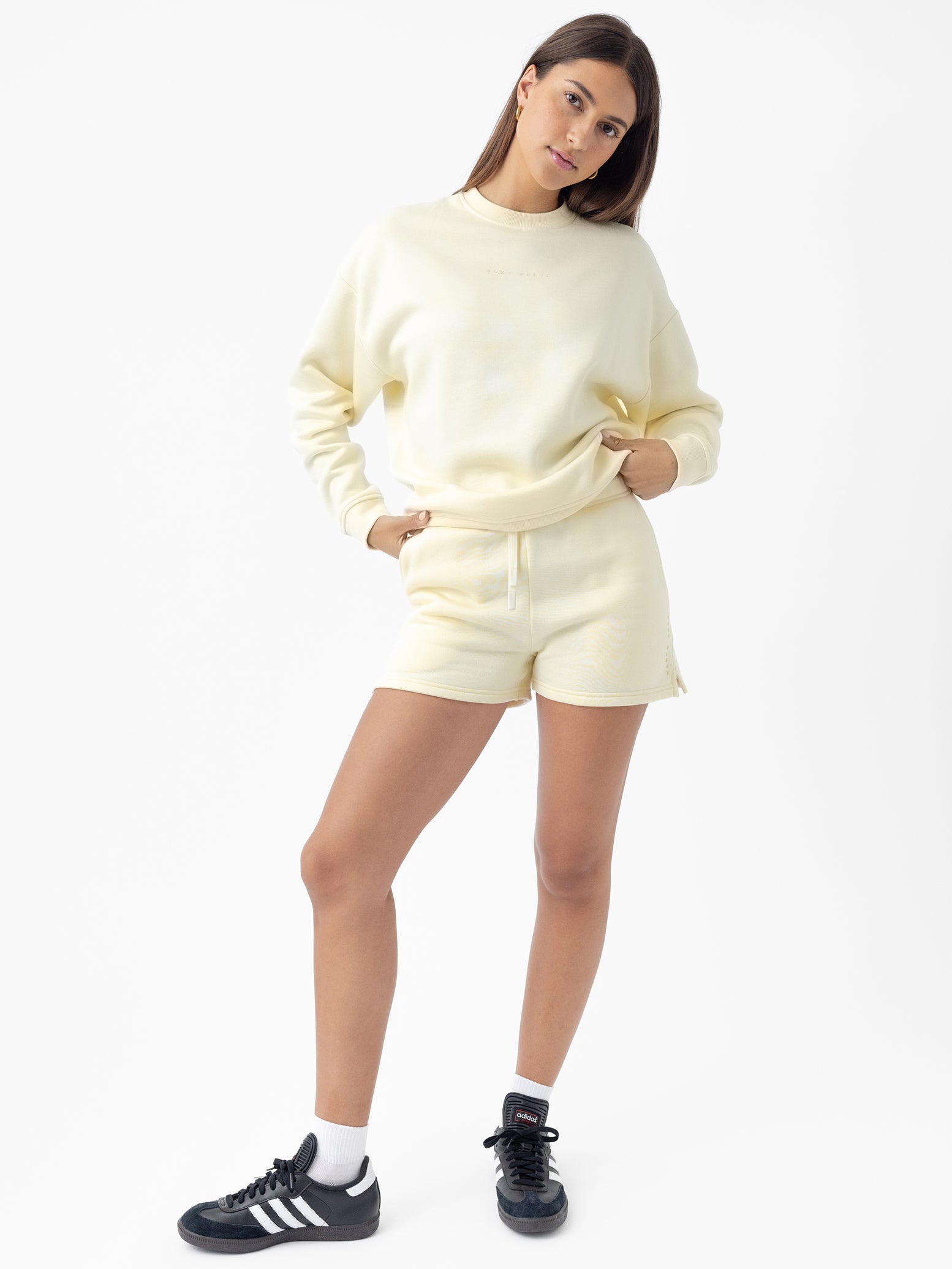 A woman with long hair stands in a relaxed pose wearing Cozy Earth's Women's CityScape Crewneck in light yellow along with matching shorts. She finishes the look with black sneakers and white socks against a plain white background. 