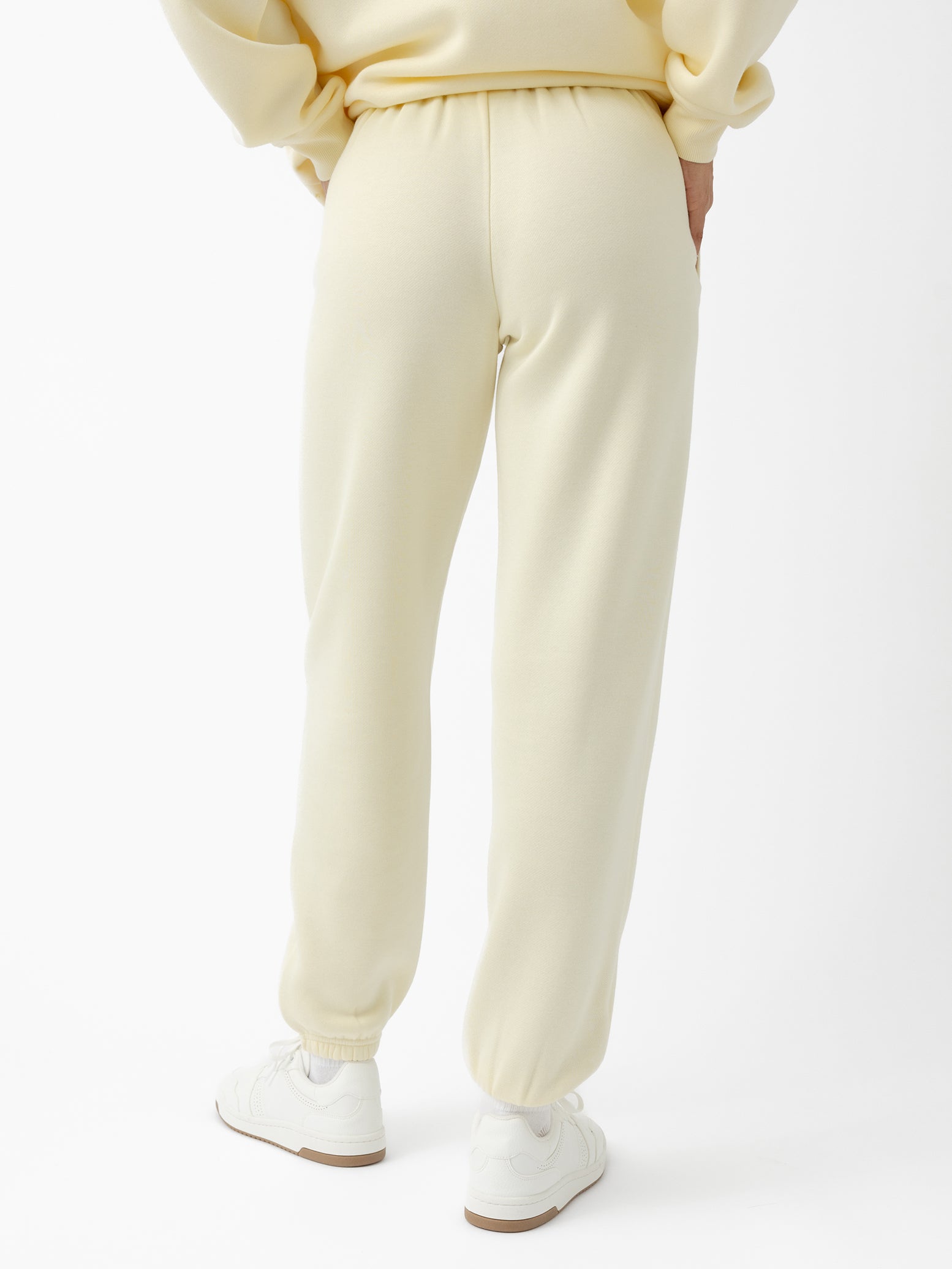 Rear view of a person wearing Women's CityScape Sweatpant by Cozy Earth in light yellow, paired with a matching sweatshirt. The sweatpants have an elastic waistband and cuffs, complemented by white sneakers against a plain white background. 