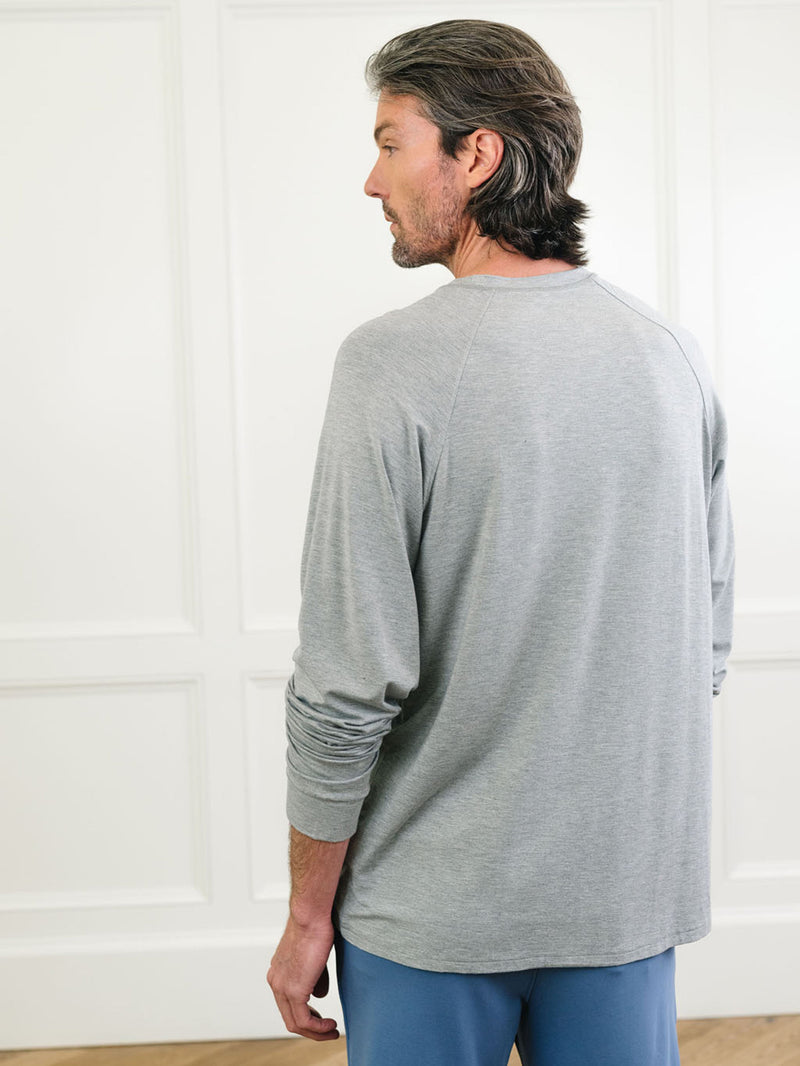 A man with medium-length hair stands facing away, wearing a long-sleeve, light gray shirt and blue pants from the Men's Stretch-Knit Long Sleeve Bamboo Pajama Set by Cozy Earth. The background features white paneled walls and a beige floor.