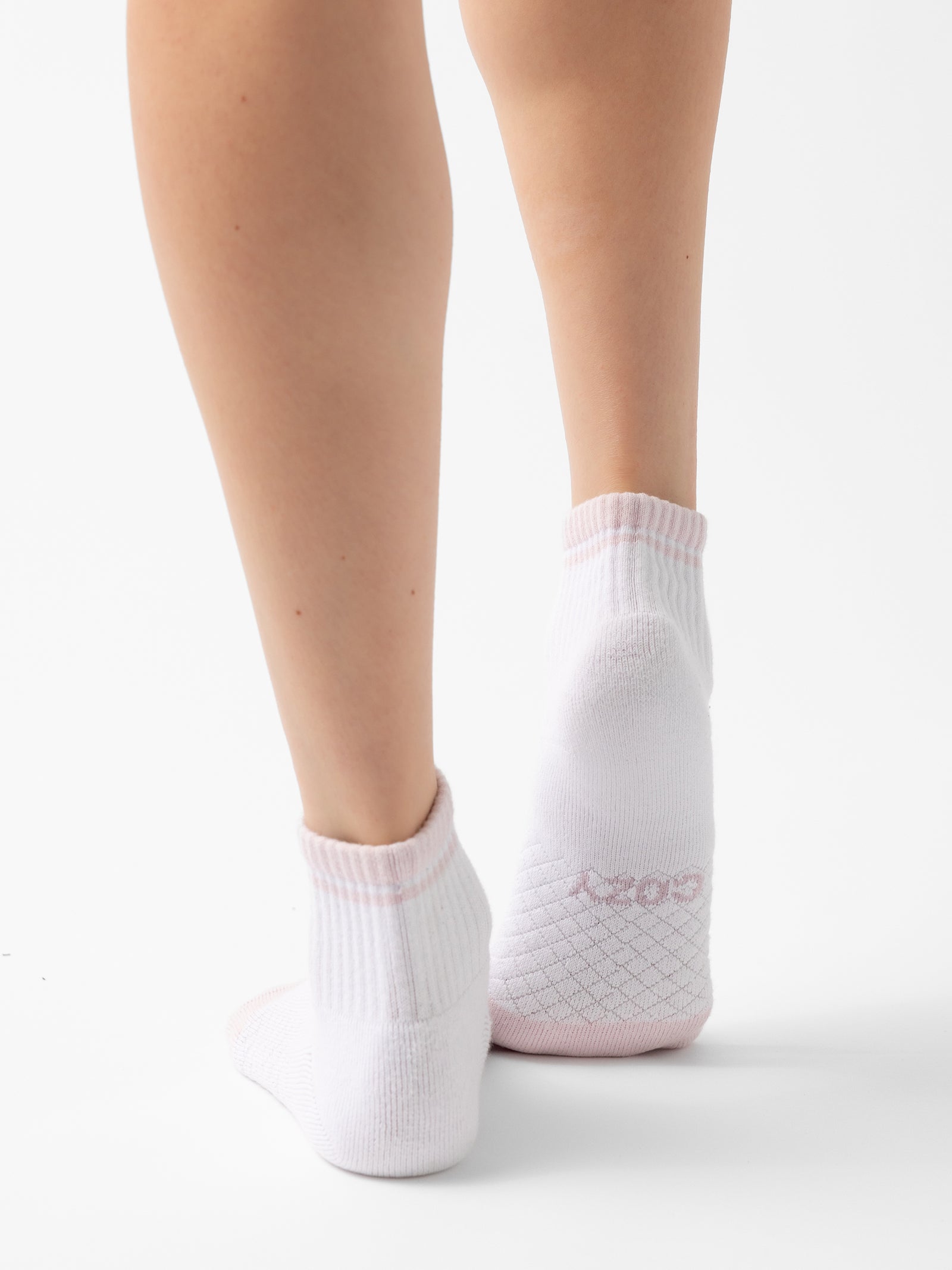 A close-up view of a person wearing a pair of white ankle socks with light pink trim from the Essential Quarter Sock 2-Pack by Cozy Earth. The image shows the back of the legs and feet, highlighting the detailed knit pattern on the heels and the word "Cozy" on the socks' cuffs. 