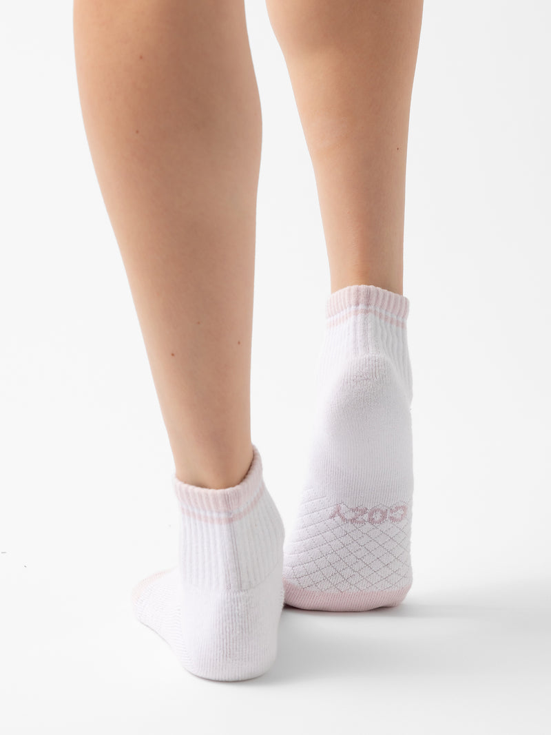 A close-up view of a person wearing a pair of white ankle socks with light pink trim from the Essential Quarter Sock 2-Pack by Cozy Earth. The image shows the back of the legs and feet, highlighting the detailed knit pattern on the heels and the word "Cozy" on the socks' cuffs.