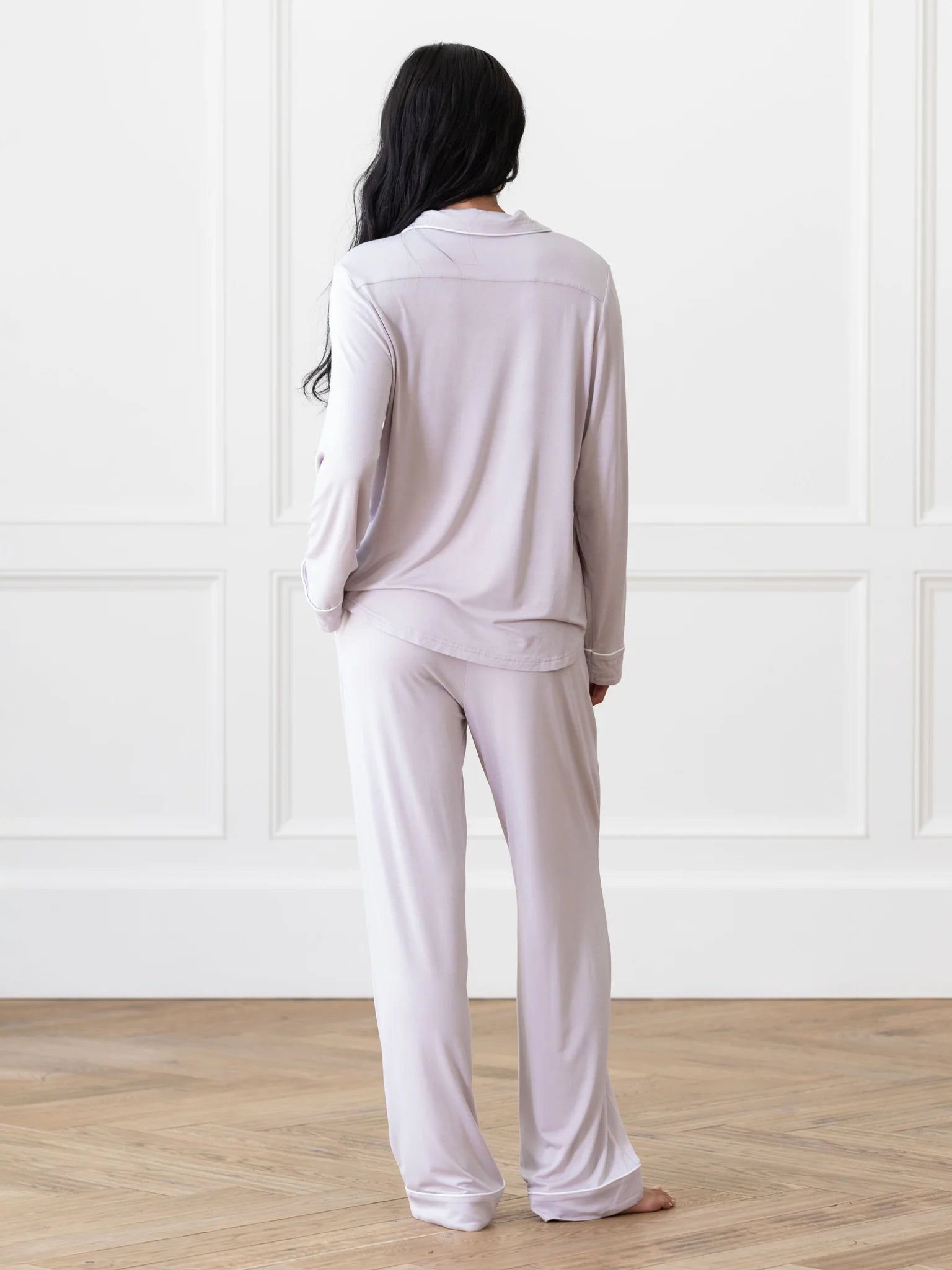 A person with long dark hair stands barefoot in a bright room facing a white paneled wall, wearing the light gray Bedtime Bundle pajama set from Cozy Earth.