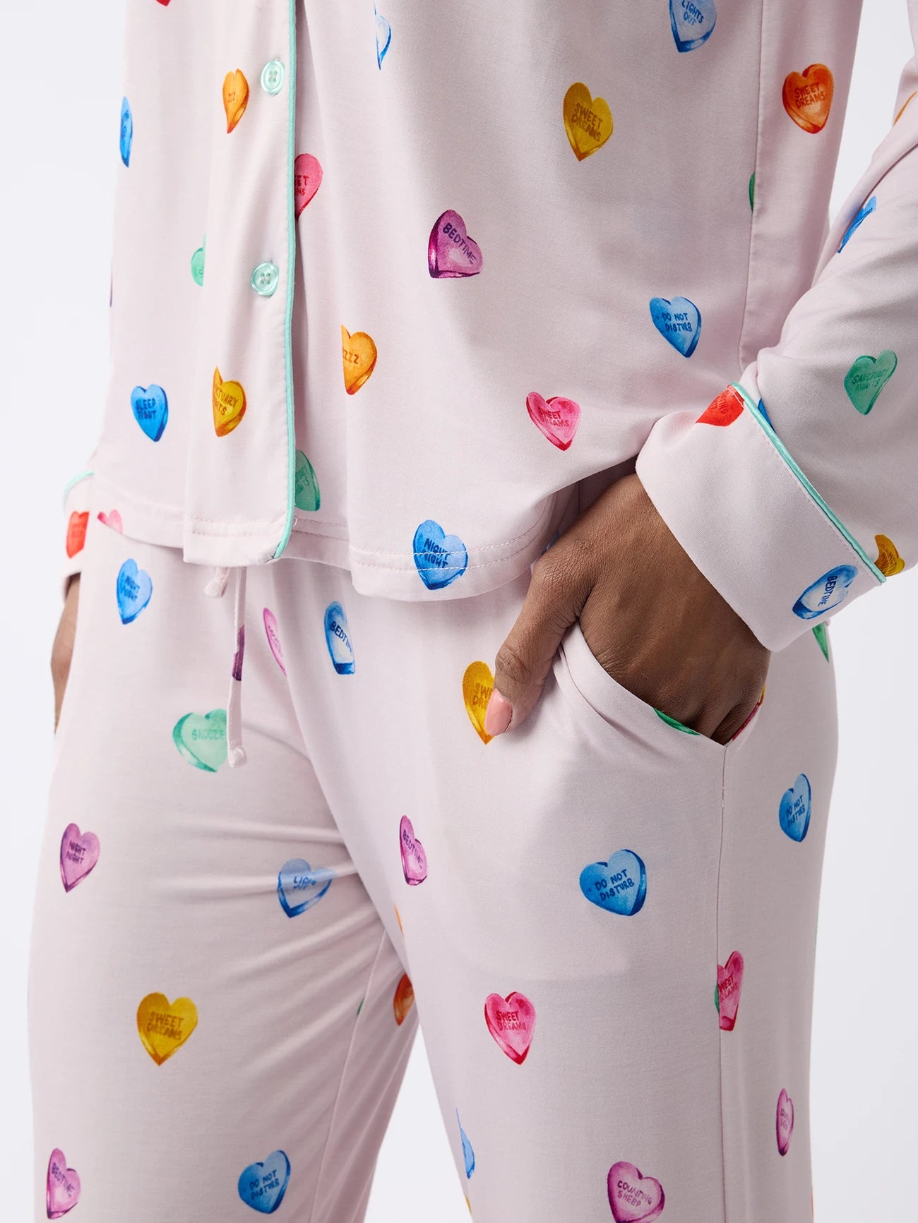 A person in pastel purple Women's Stretch-Knit Bamboo Pajama Pant by Cozy Earth, featuring colorful heart patterns with small text. The pants have buttons and pockets, with a hand resting in one pocket. The backdrop is plain white. 