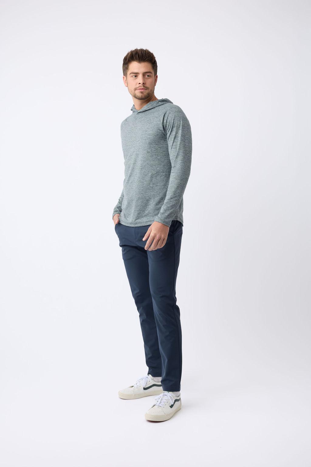 Against a plain white background, a man with short hair stands wearing Cozy Earth's Men's Lightweight Performance Hoodie in gray, paired with dark blue pants and white sneakers. His hands rest in his pockets as he gazes slightly to the left. |Color:Heather Dawn