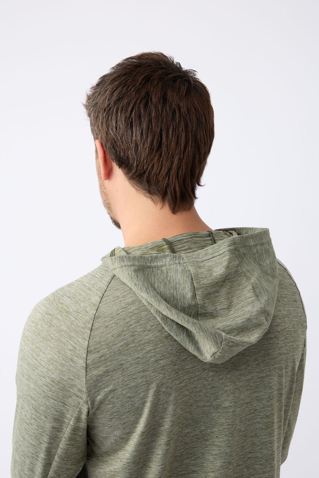 A person with brown hair is seen from behind wearing a Cozy Earth Men's Lightweight Performance Hoodie in light green heather. The background is plain and light-colored, creating a neutral setting.