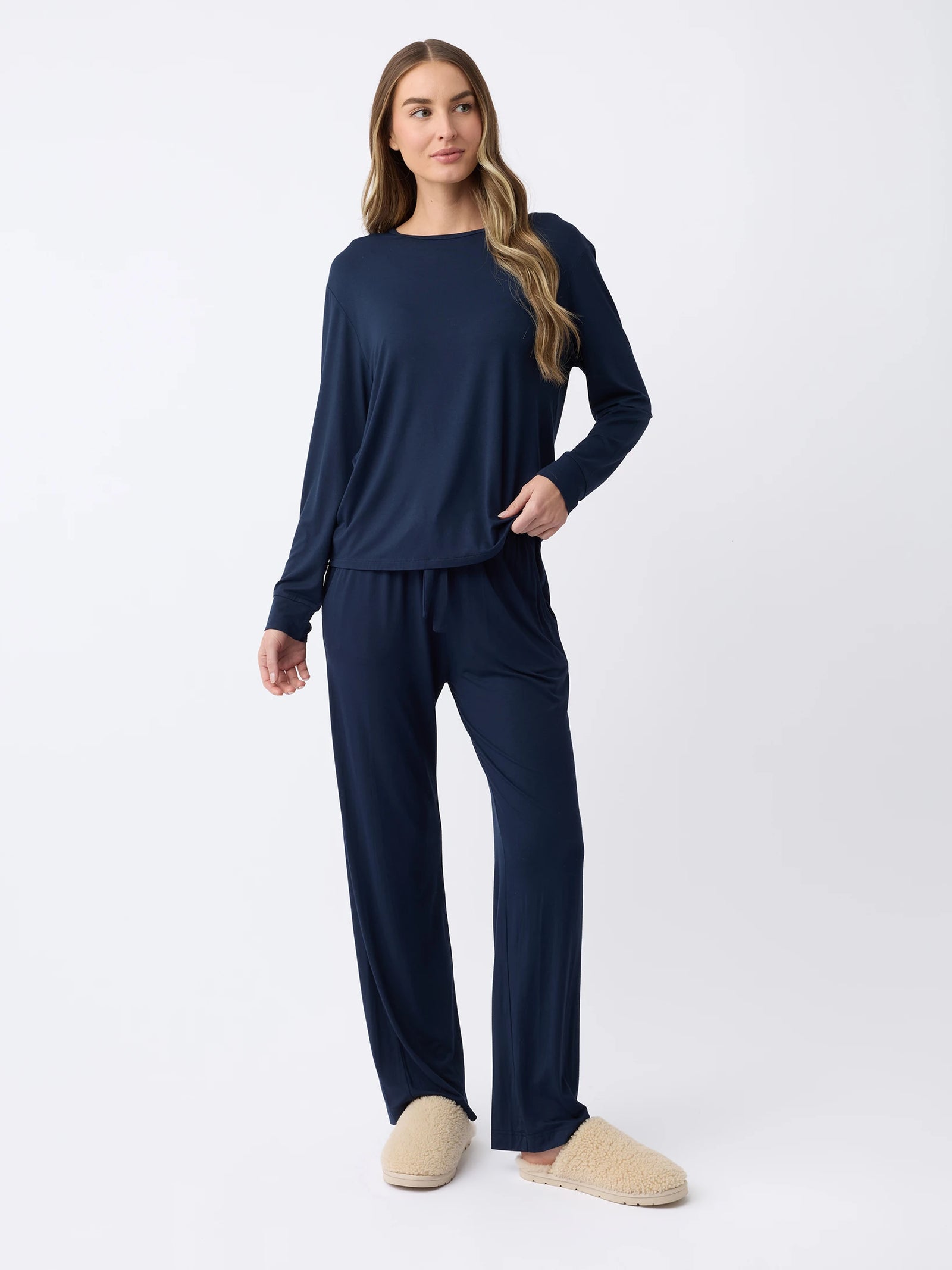 Against a plain background, a woman with long, wavy hair wears the Cozy Earth Women's Stretch Knit Bamboo Long Sleeve Lounge Tee in dark blue, paired with matching pants and light beige slippers, with one hand in a pocket. 