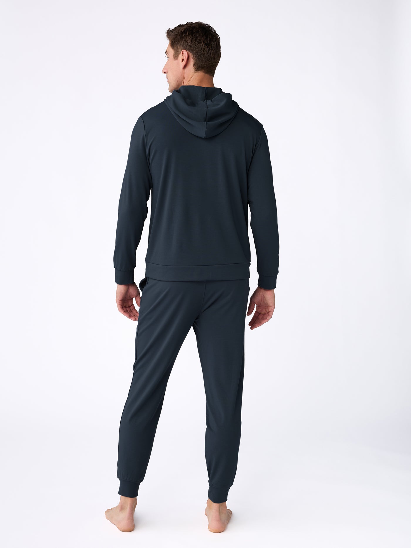 A man stands barefoot, facing away from the camera, wearing the Cozy Earth Men's Brushed Bamboo Hoodie & Jogger Set in dark color against a plain white background. |Color:Navy