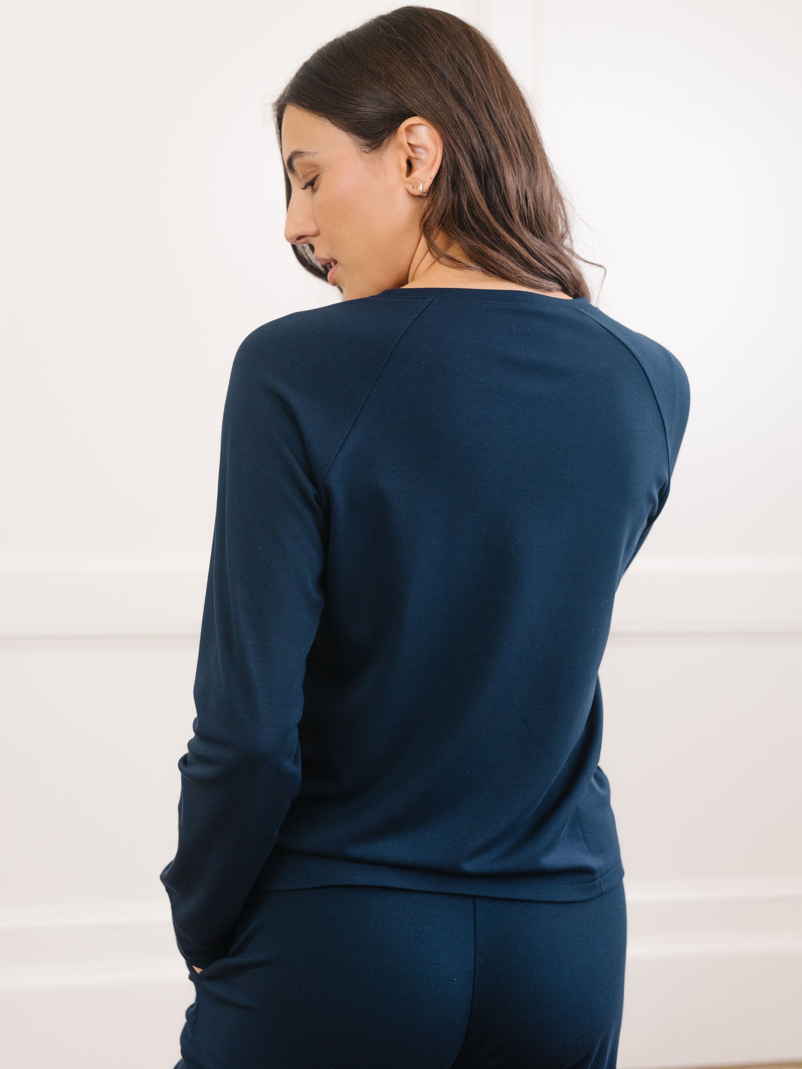 A woman with long dark hair faces away from the camera, wearing Cozy Earth's navy Women's Ultra-Soft Bamboo Wide Leg Pull On Pant & Pullover Crew Set. She stands in a room with a white wall, appearing relaxed. 