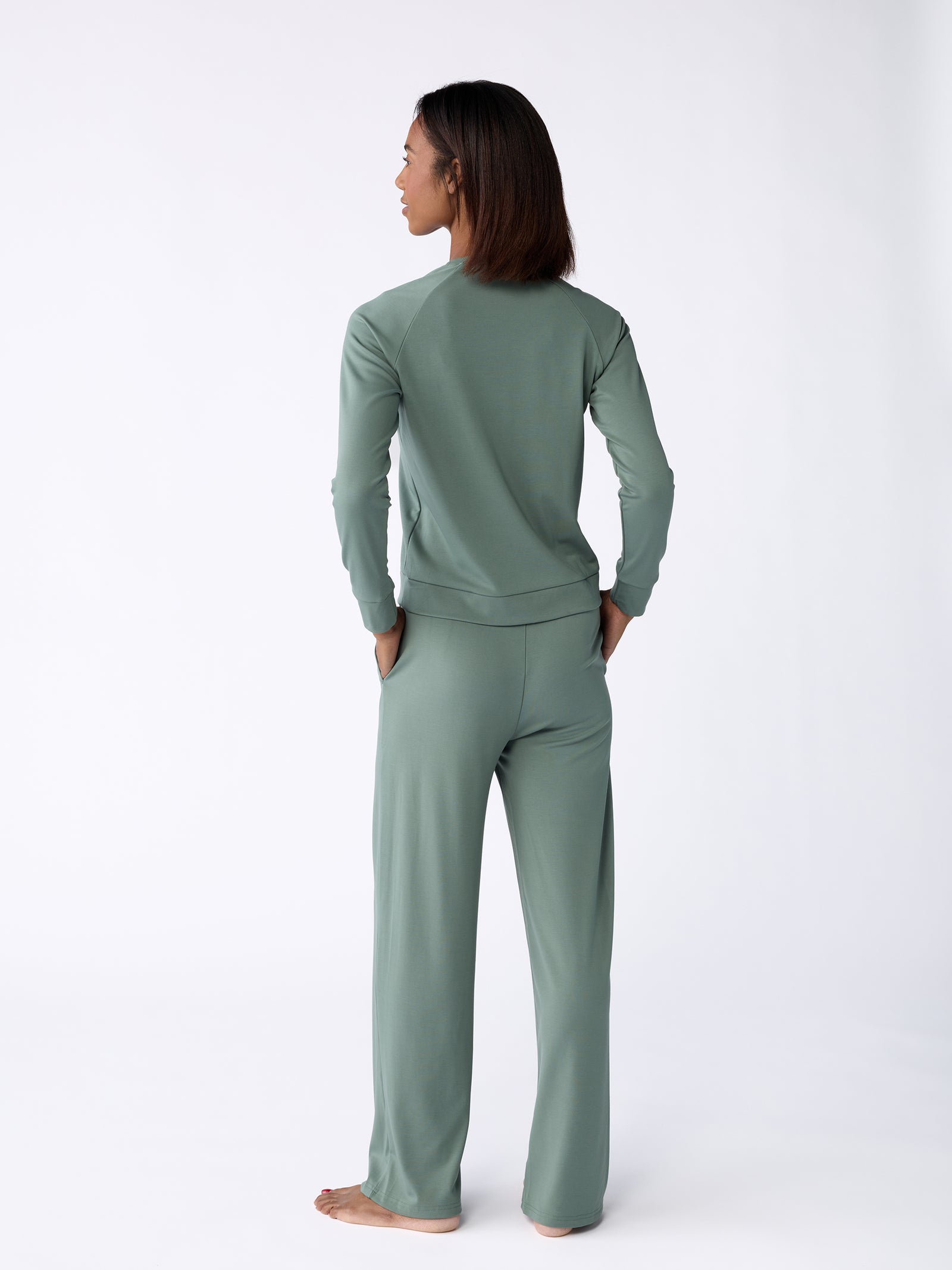 A person wearing the Women's Ultra-Soft Bamboo Pullover Crew by Cozy Earth in sage green stands barefoot on a plain white background. They have their back turned, with a relaxed posture and hands casually tucked into their pockets. 