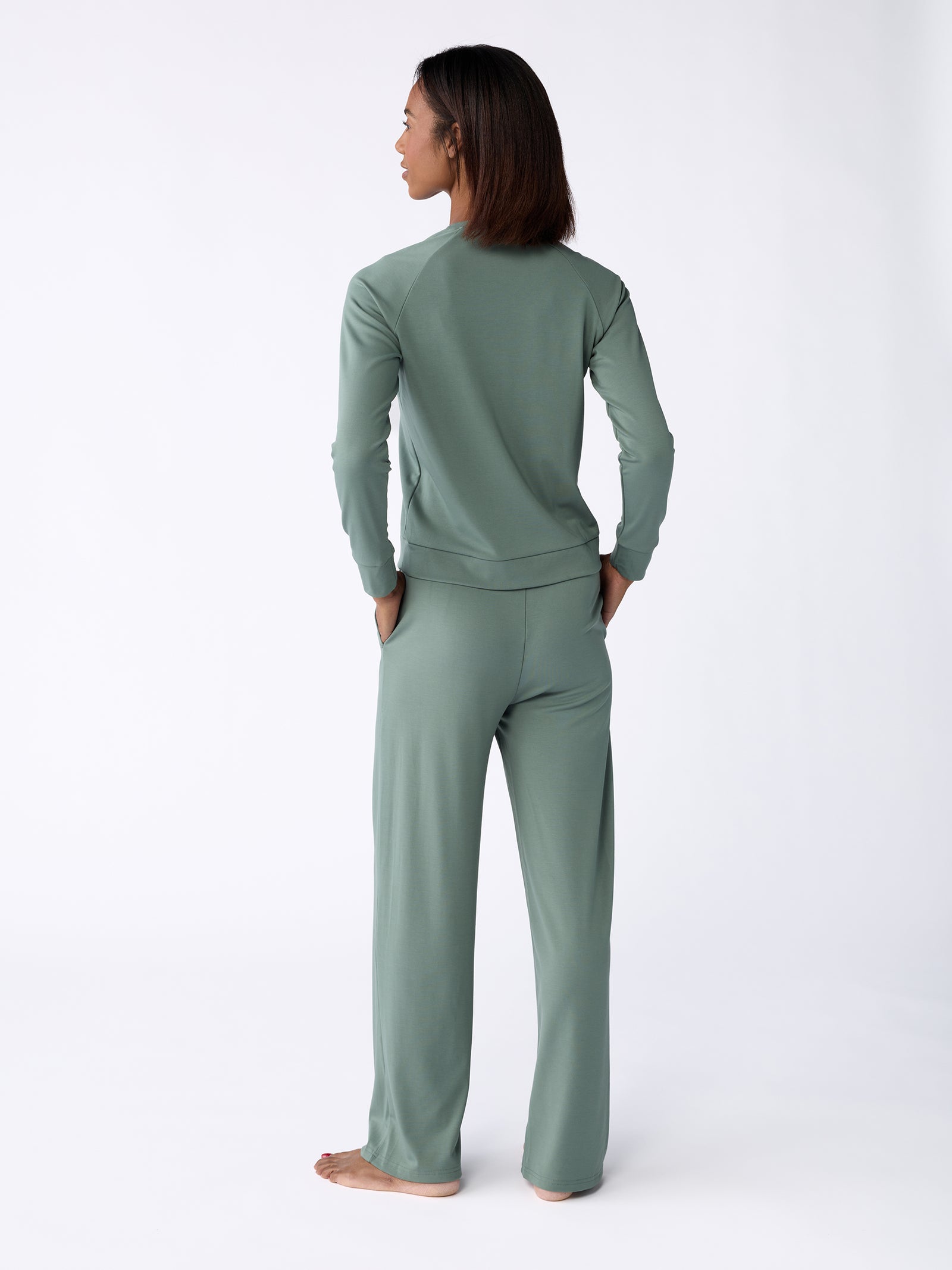 A person stands barefoot on a white background, wearing a sage green long-sleeve top paired with Cozy Earth's Women's Ultra-Soft Bamboo Wide Leg Pull On Pant. They are facing away, looking to the side. The outfit appears comfortable and casual. 