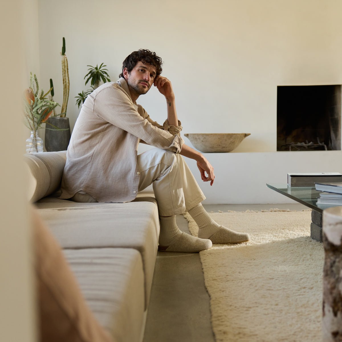 A man with curly hair in a light beige shirt, cream pants, and socks sits on a minimalist beige sofa in a neutral-toned living room. A cactus, other plants, a fireplace, and a glass coffee table are in the background. He's wearing Cozy Earth's Lakehouse Clog shoes. 