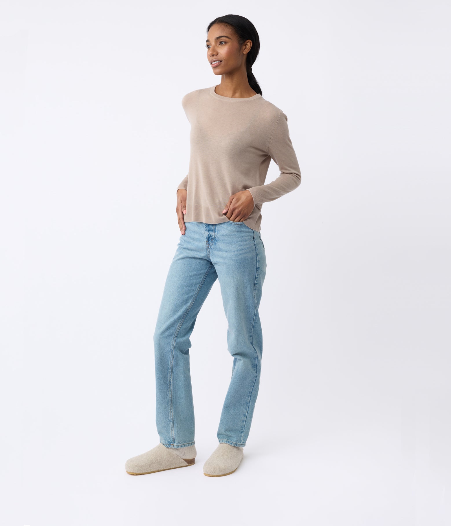 A person wearing a beige sweater, light blue jeans, and Cozy Earth's Lakehouse Clog stands against a plain white background, smiling with hands on hips. 