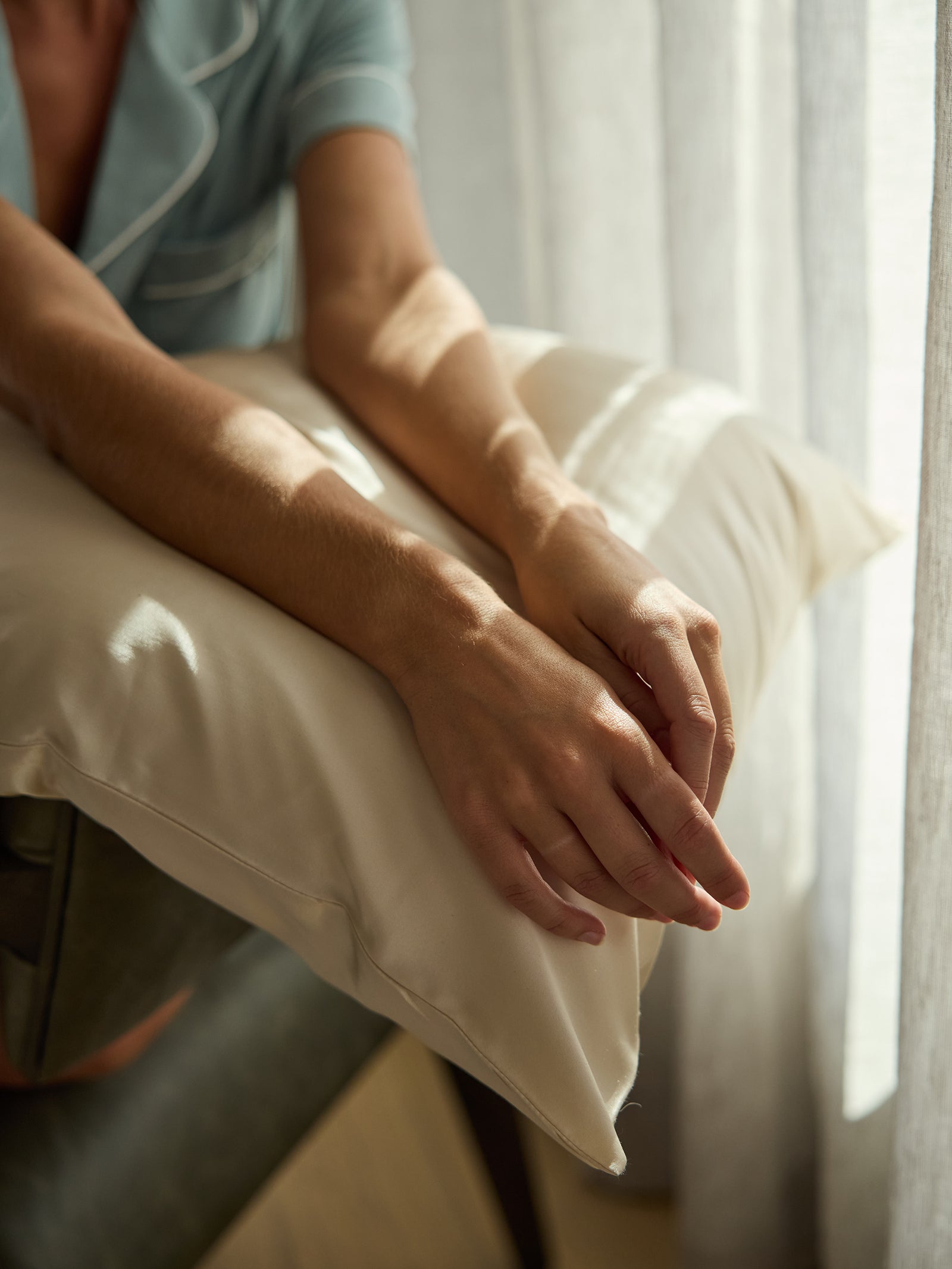 A person wearing a light-colored robe rests their arms on a Cozy Earth Silk Pillowcase by a window with sheer curtains. Soft sunlight gently illuminates the scene, creating a calm and peaceful atmosphere. 