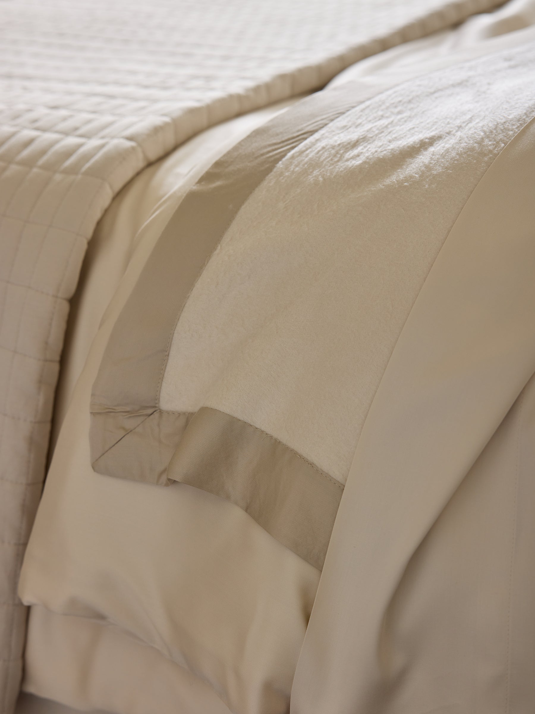 A close-up of a neatly made bed featuring beige and cream-colored bedding incorporates the luxurious Bamboo Blanket from Cozy Earth. The comforter is lightly quilted, and on top lies the soft, satin-bordered Bamboo Blanket, smoothly laid out. The textures suggest a comfortable and inviting sleeping space. |Color:Oat/Driftwood
