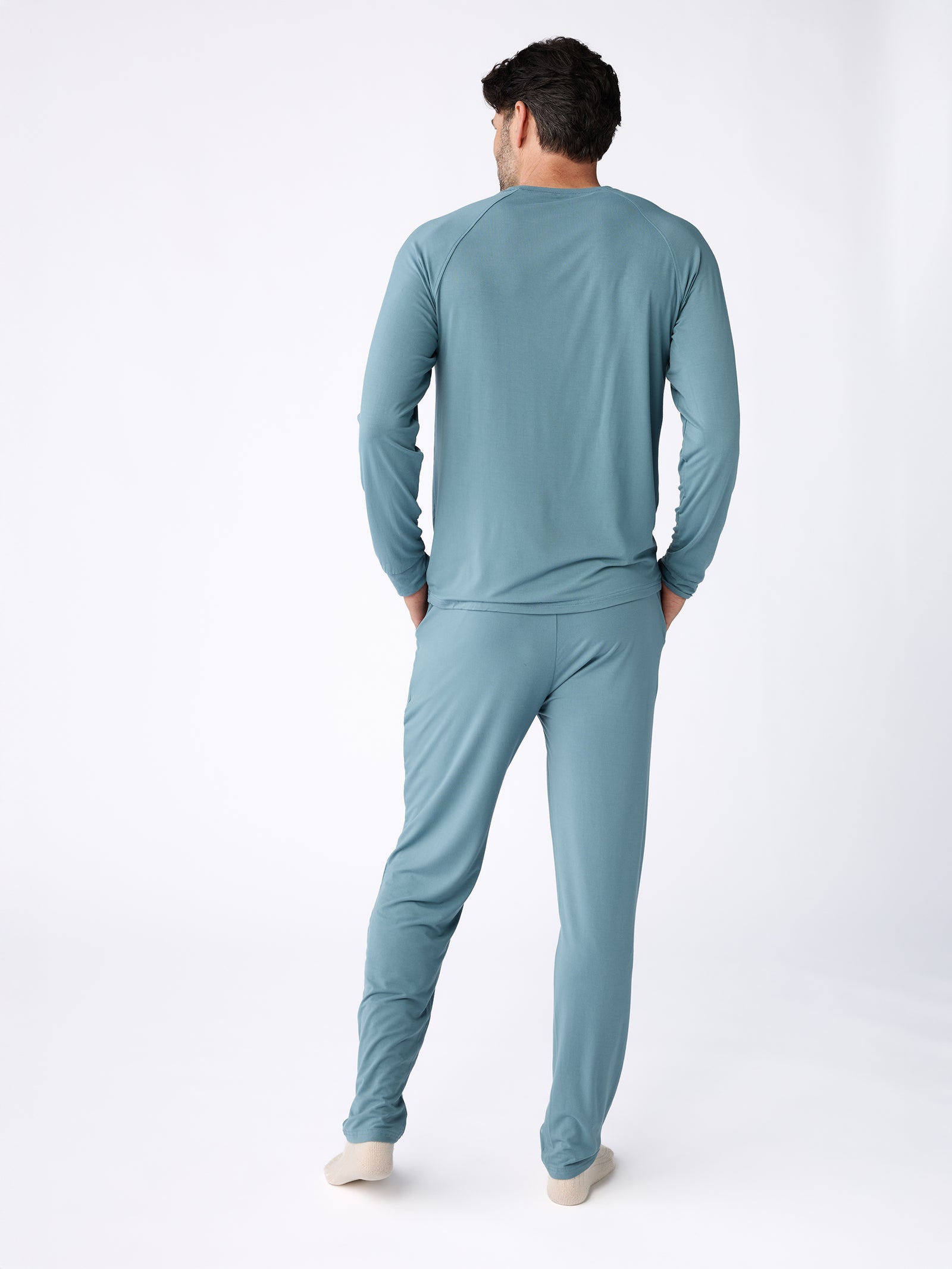 A man is seen from behind in Cozy Earth's teal Men's Stretch-Knit Bamboo Long Sleeve pajamas, which include a long-sleeve top and matching pants. The background is a simple white studio setting. 