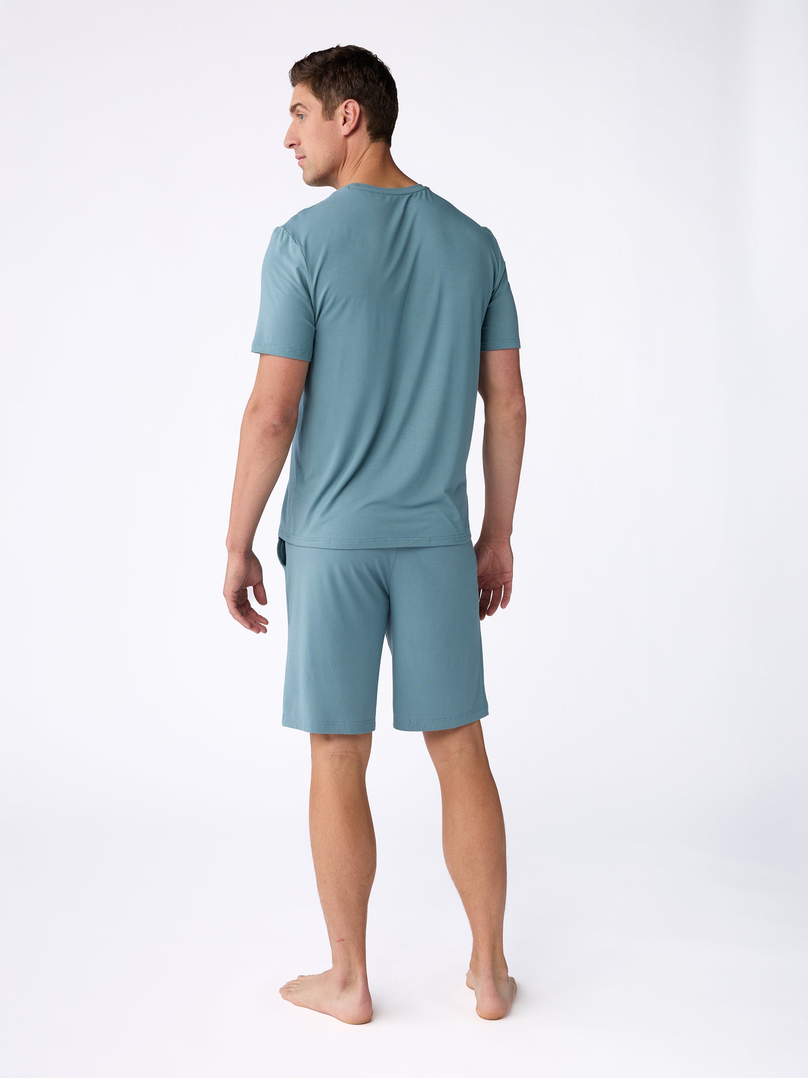 A man is standing barefoot on a white background, wearing a matching set of Cozy Earth's Men's Stretch-Knit Bamboo Pajama Shorts and T-shirt in blue-green. He is facing away from the camera, with his head turned slightly to the left. 