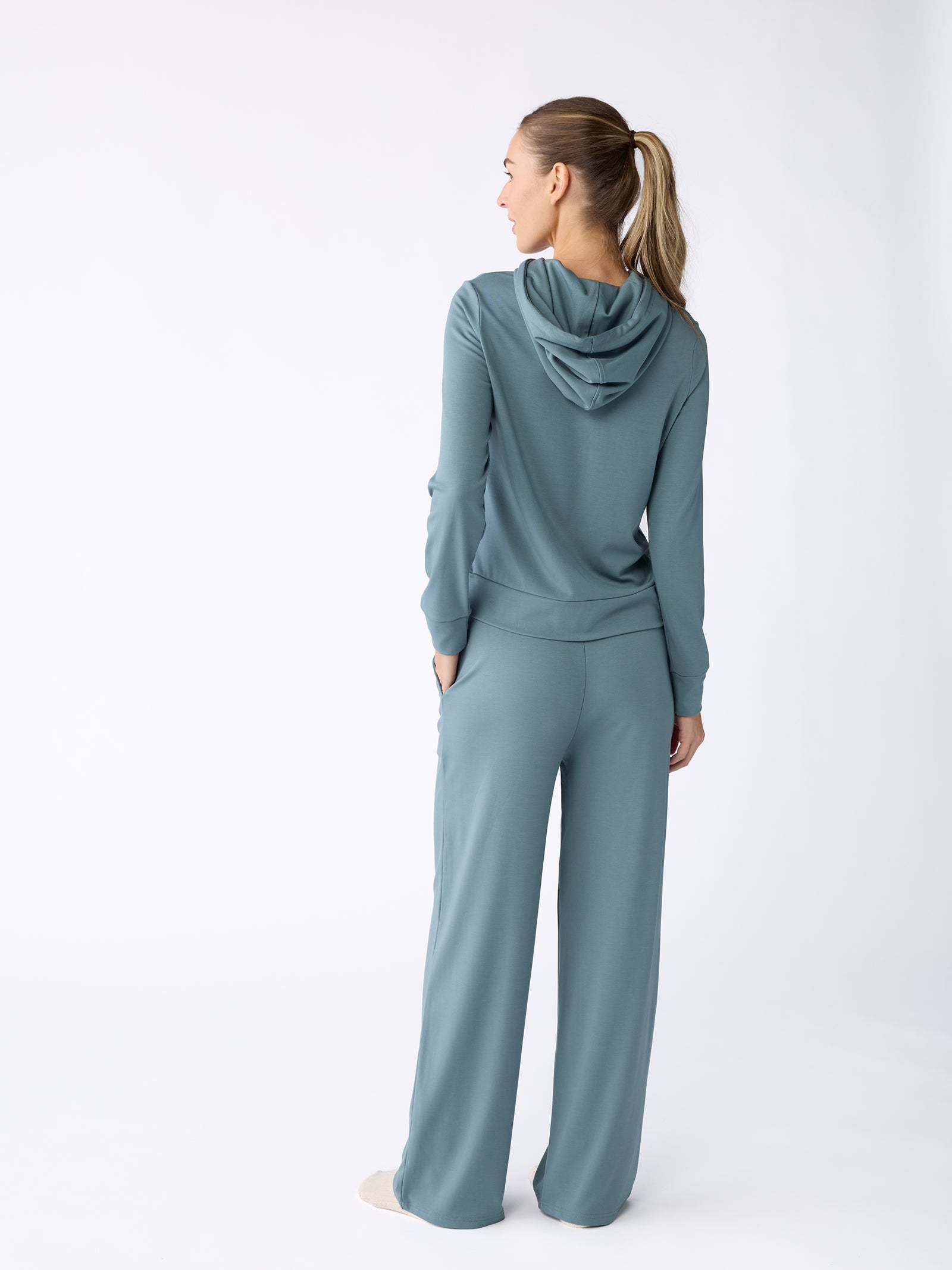 A person wearing the Women's Ultra-Soft Bamboo Wide Leg Pull On Pant & Hoodie Set from Cozy Earth, in teal, stands facing away on a plain background. They have long hair in a ponytail and one hand in their pocket. 