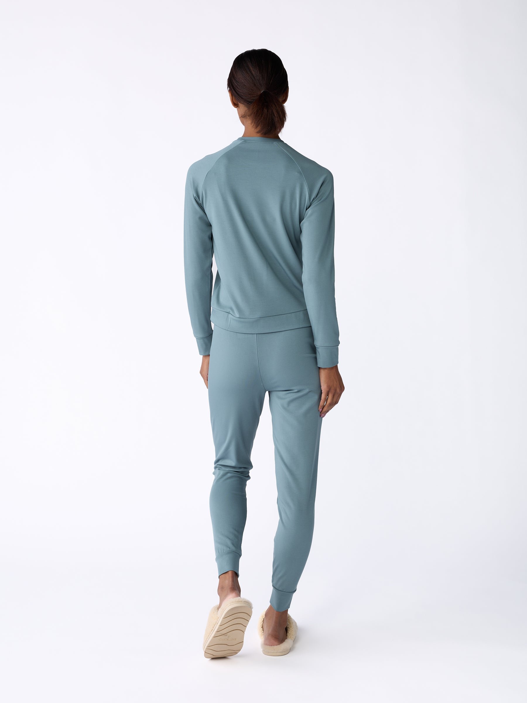 A woman is seen facing away, dressed in a matching blue-green tracksuit, featuring the Women's Bamboo Jogger Pant by Cozy Earth, complemented by beige slip-on shoes. The plain white background highlights the clothing ensemble. 