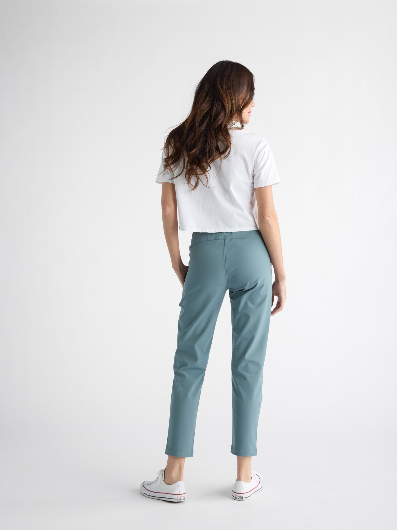 A woman with long hair stands in a minimalist, light-gray studio, facing away. She wears a white t-shirt, white sneakers, and the Women's Always Cropped Pant by Cozy Earth in teal. 