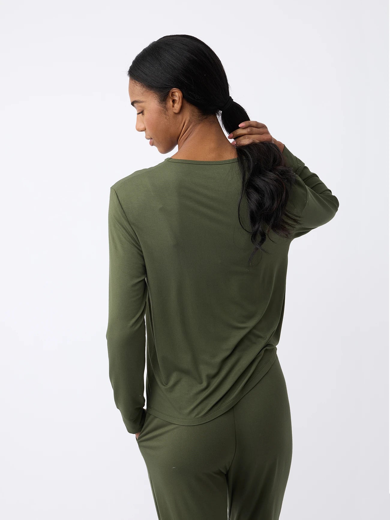 A woman with long dark hair, facing away, is wearing Cozy Earth's Women's Stretch Knit Bamboo Long Sleeve Lounge Tee in olive green with matching pants. Their right hand touches their hair against a plain white wall backdrop. 