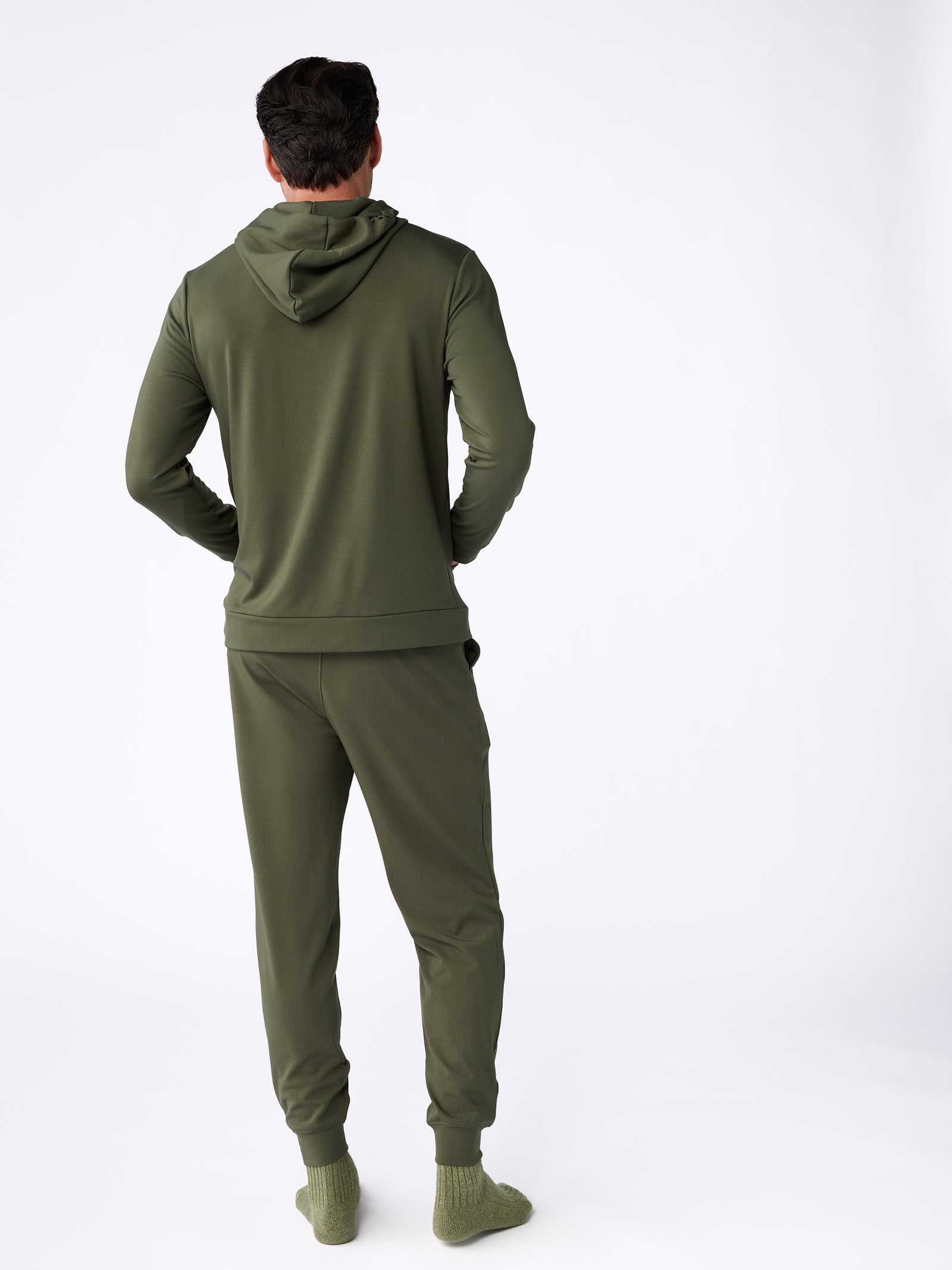 A person is shown from behind against a stark white backdrop, dressed in the Men's Ultra-Soft Bamboo Hoodie and corresponding joggers in a shade of olive green. Their outfit is completed with matching green socks. 