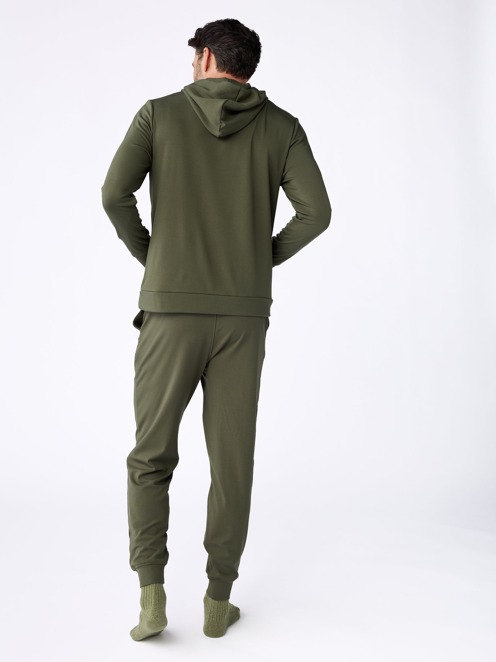 A person is modeling the Cozy Earth Men's Ultra-Soft Bamboo Pullover Crew and matching sweatpants in olive green, with their back to the camera. They are also wearing green socks in a plain white studio setting. 