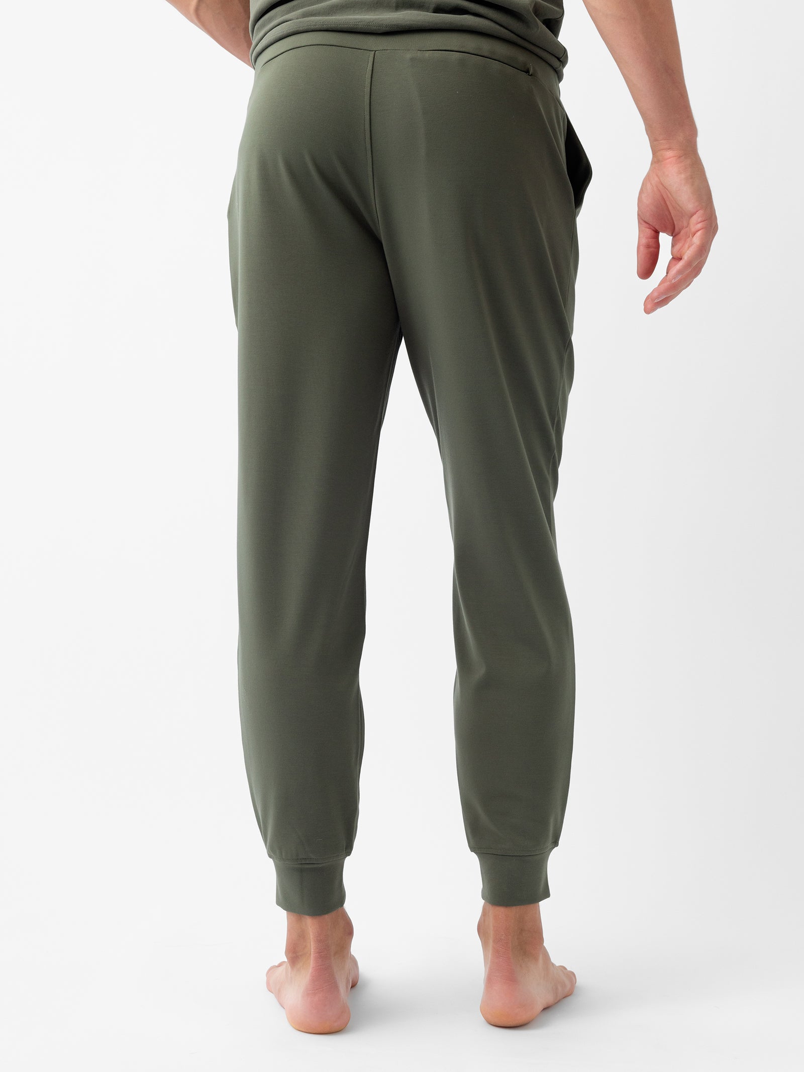 From the back, a person standing barefoot on a white surface is shown wearing olive green Men's Ultra-Soft Bamboo Jogger Pants by Cozy Earth and a matching green top. The pants feature a relaxed fit with cuffed hems, and one hand is partially visible at their side. 