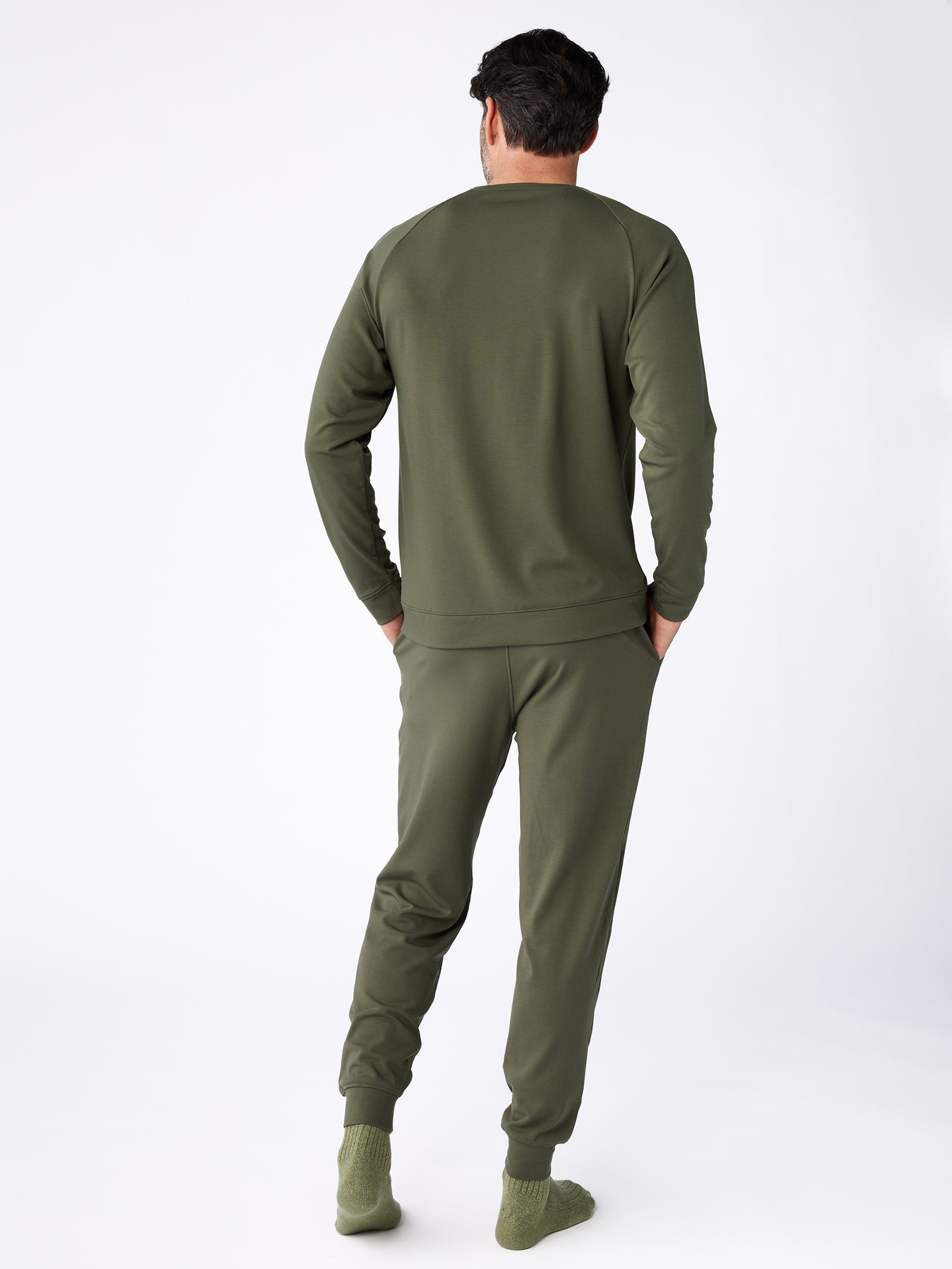 A man is wearing a Cozy Earth Men's Ultra-Soft Bamboo Pullover Crew in a matching olive green color, paired with pants. His outfit includes the long-sleeve top and similarly colored socks as he stands with his back to the camera against a plain white background. 