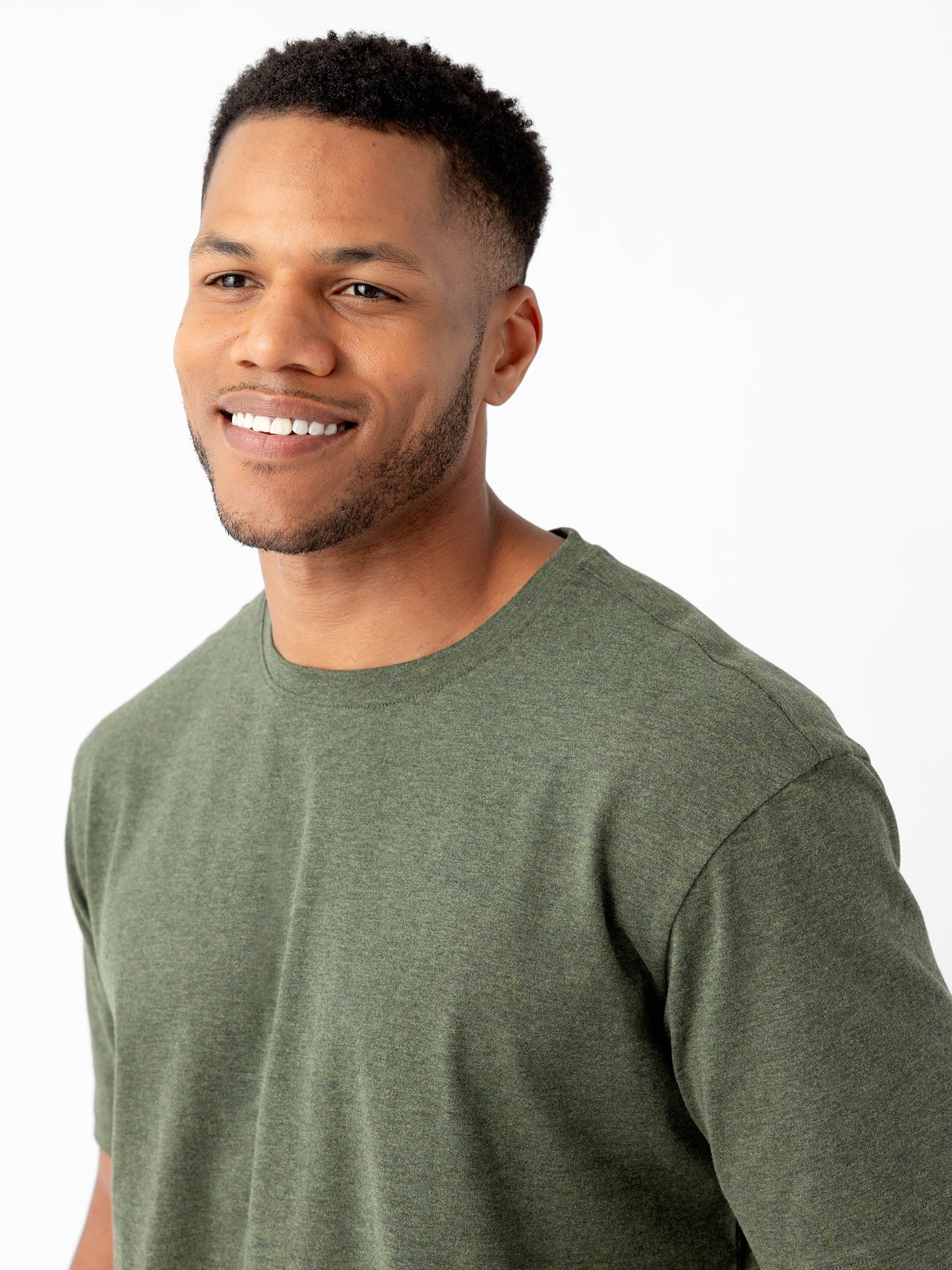 A man with short, dark hair and a beard smiles at the camera. He is wearing a Men's All Day Tee by Cozy Earth in green and stands against a plain white background. 