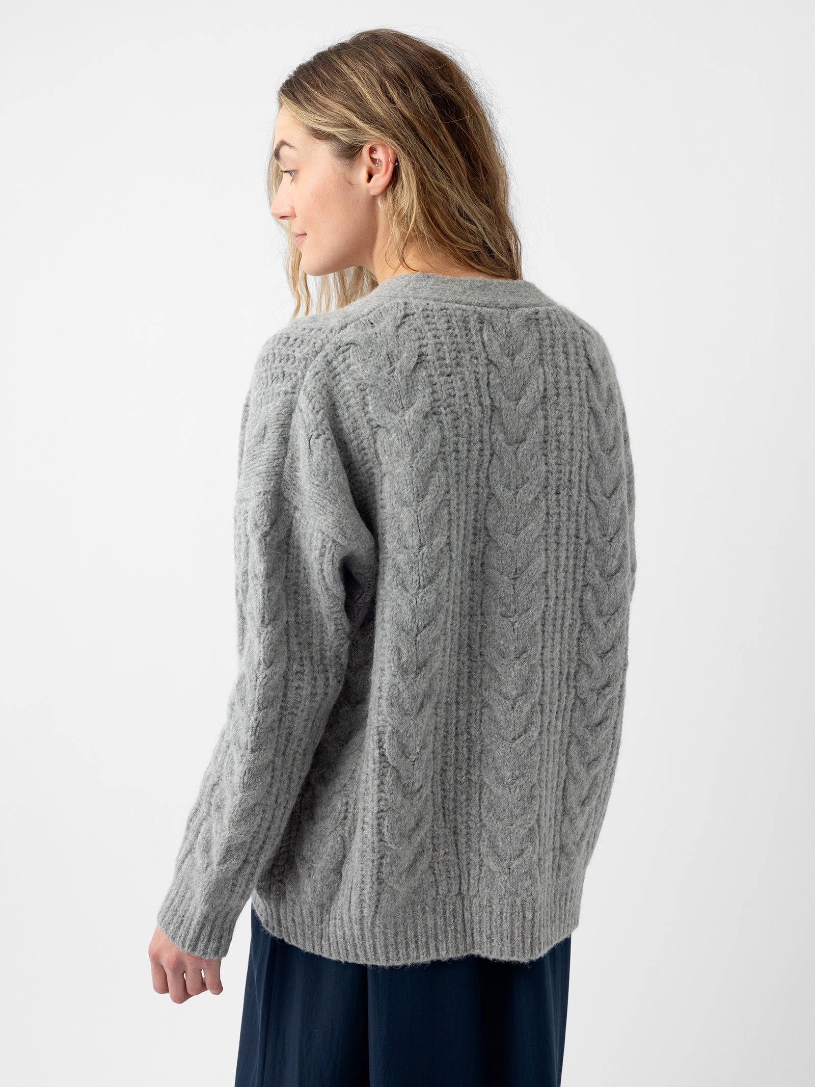 A woman with long hair is wearing a gray Oversized Cable Knit Cardigan from Cozy Earth. She is facing away, with her head turned slightly to the side, against a plain white background. The bottom of her skirt peeks into view at the edge of the image. 