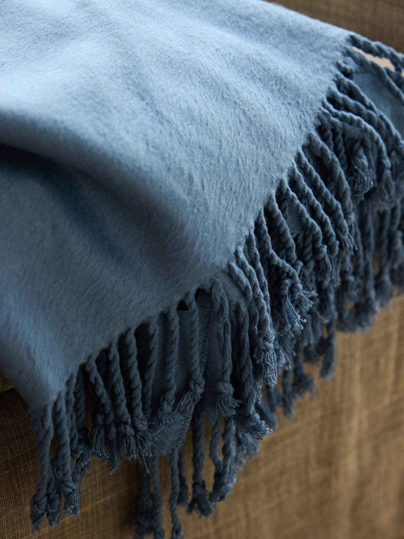 A close-up of a Cozy Earth Bamboo Tassel Throw in blue, draped over the arm of a beige couch. The throw has a soft texture and twisted tassel edges that hang loosely over the side of the furniture. 