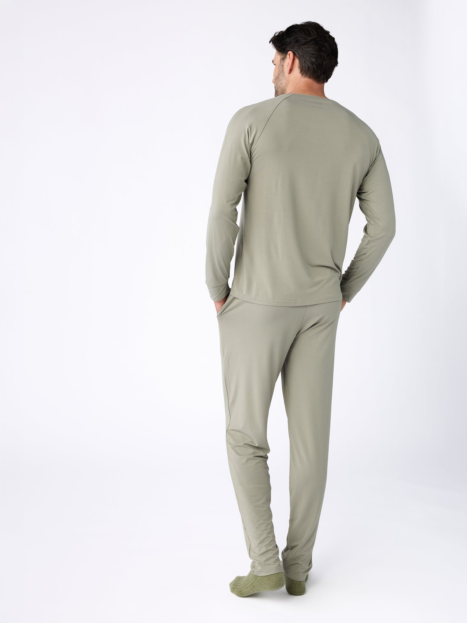 A man wearing the Men's Stretch-Knit Bamboo Long Sleeve set from Cozy Earth, consisting of a long-sleeve top and pants in olive green, stands with his back to the camera against a plain white background. 