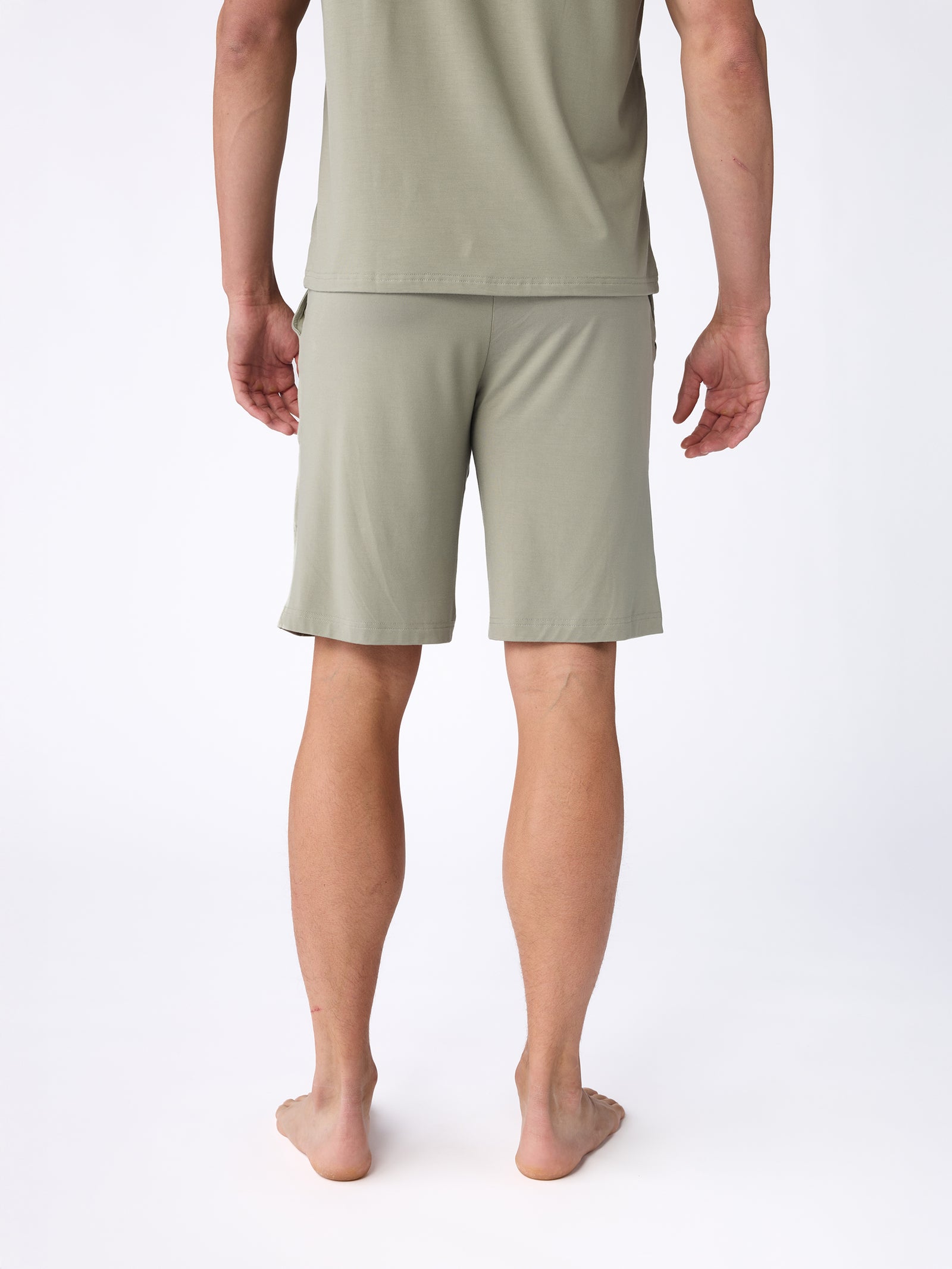 Rear view of a person standing barefoot against a white background, wearing a sage green t-shirt and matching Cozy Earth Men's Stretch-Knit Bamboo Pajama Short. The outfit appears casual and comfortable. 