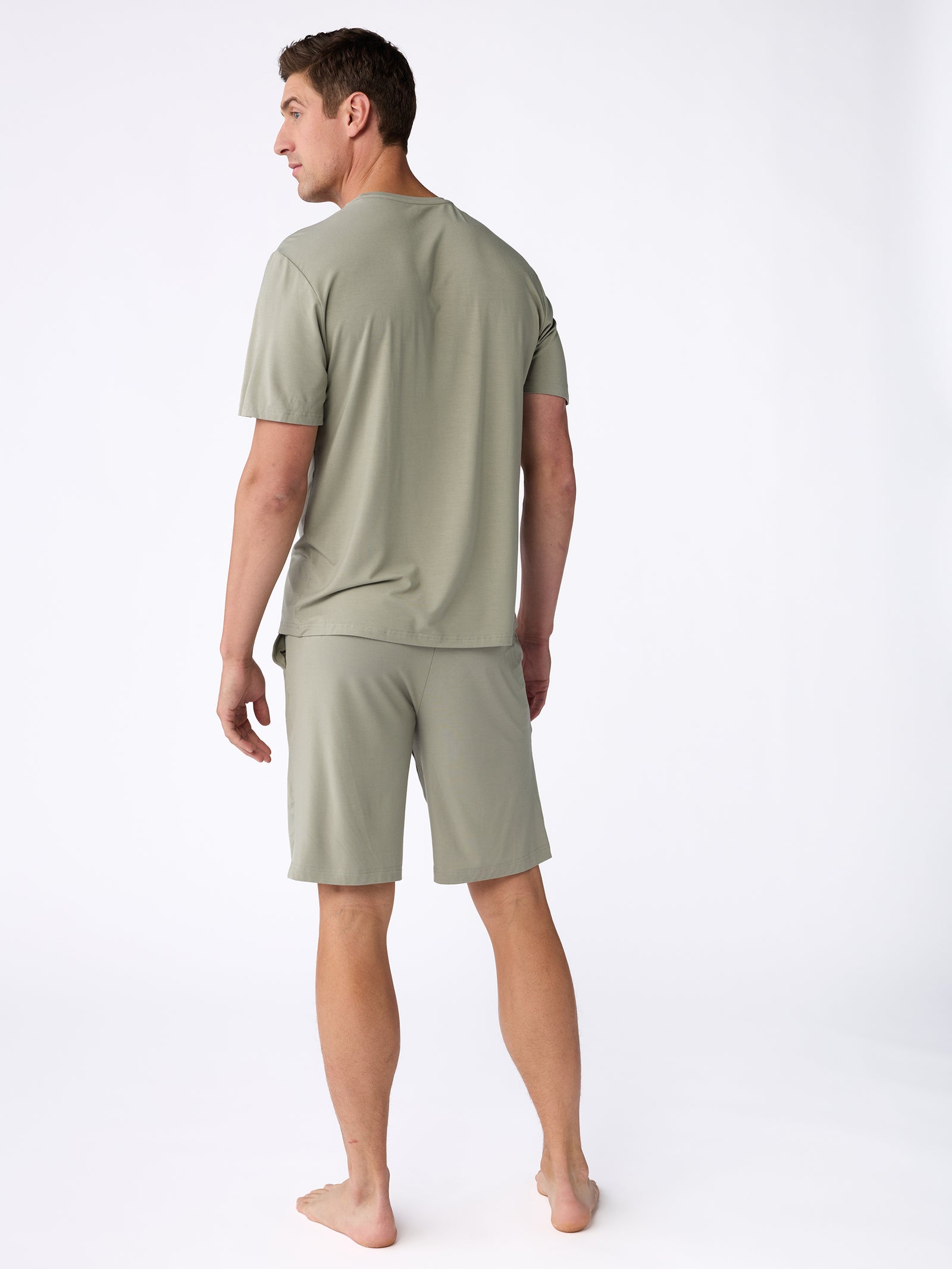 A man stands barefoot on a white background, dressed in a Cozy Earth Men's Stretch-Knit Bamboo Lounge Tee in light olive green, complemented by matching shorts. He is turned slightly away from the camera, highlighting the back and side profile of the ensemble. 