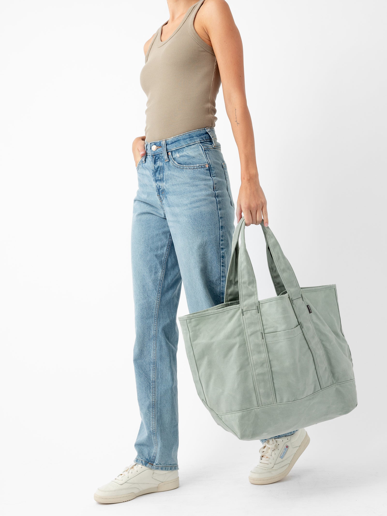 A person stands against a white background wearing a beige tank top, light blue jeans, and white sneakers. They hold a large Waxed Canvas Tote by Cozy Earth in their left hand. Their right hand is in their pocket. The outfit exudes a casual, relaxed style. 