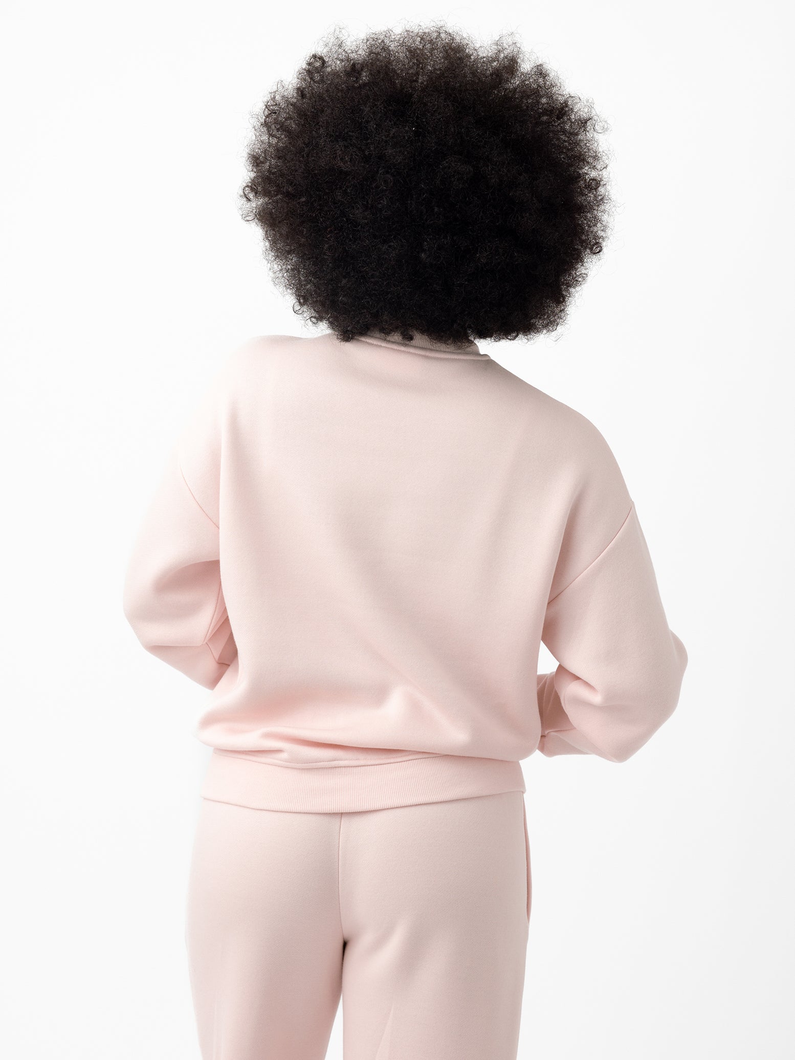 A person with curly hair is facing away, wearing a Cozy Earth Women's CityScape Crewneck in light pink with matching pants against a plain white background. 