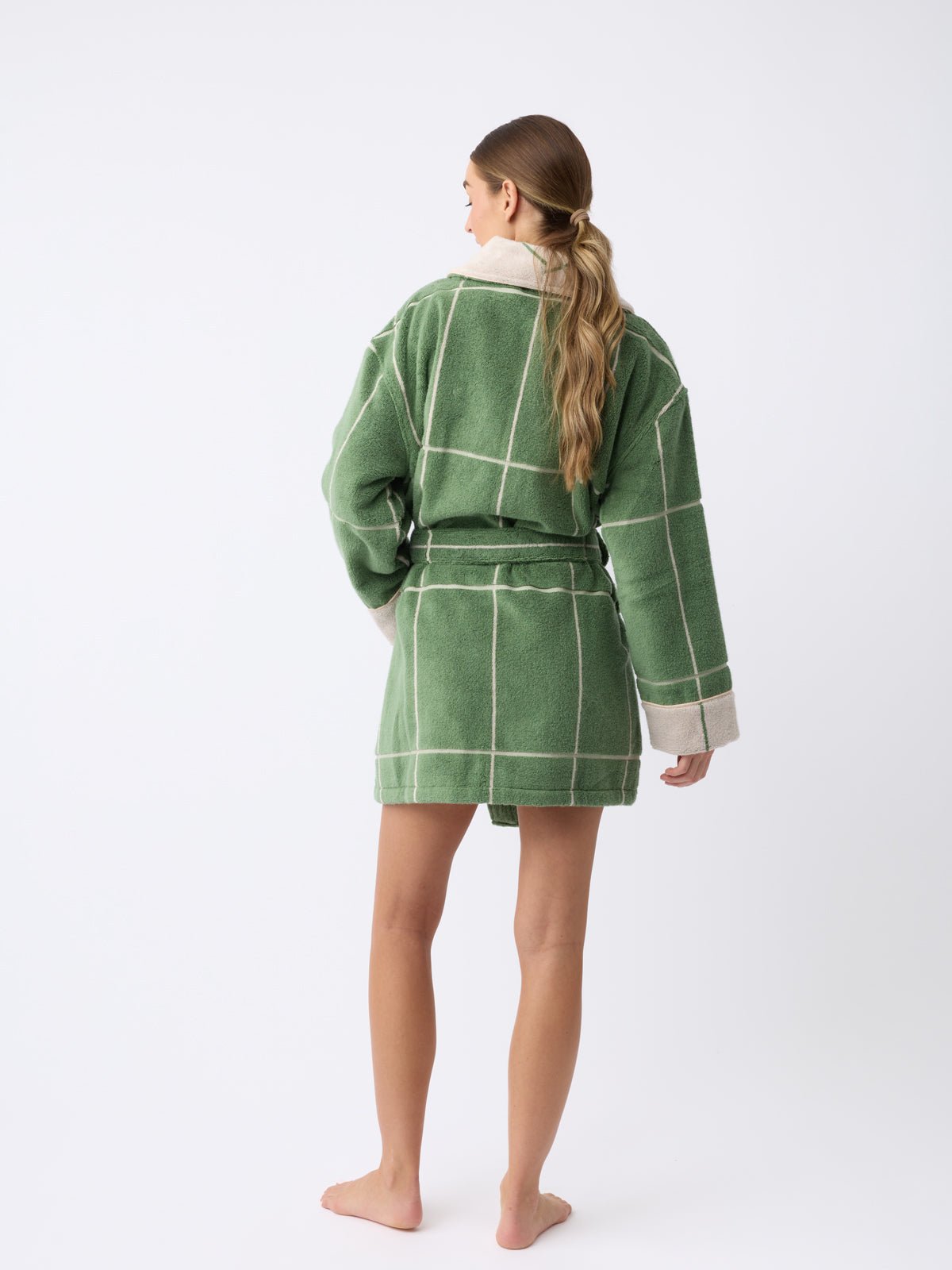 A person with long hair stands barefoot facing away, wearing a Cozy Earth Windowpane Resort Robe featuring a green plaid design with beige grid lines and a belt, set against a plain white background. 